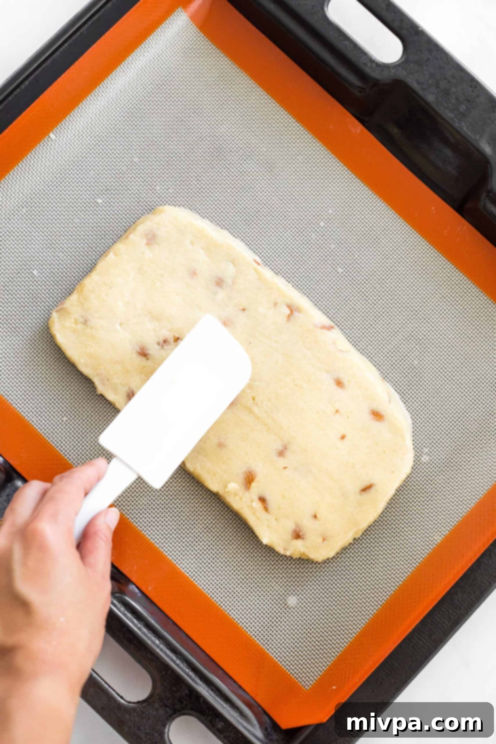 A spatula smoothing the top of a rectangular dough log on a baking sheet, preparing it for the first bake.