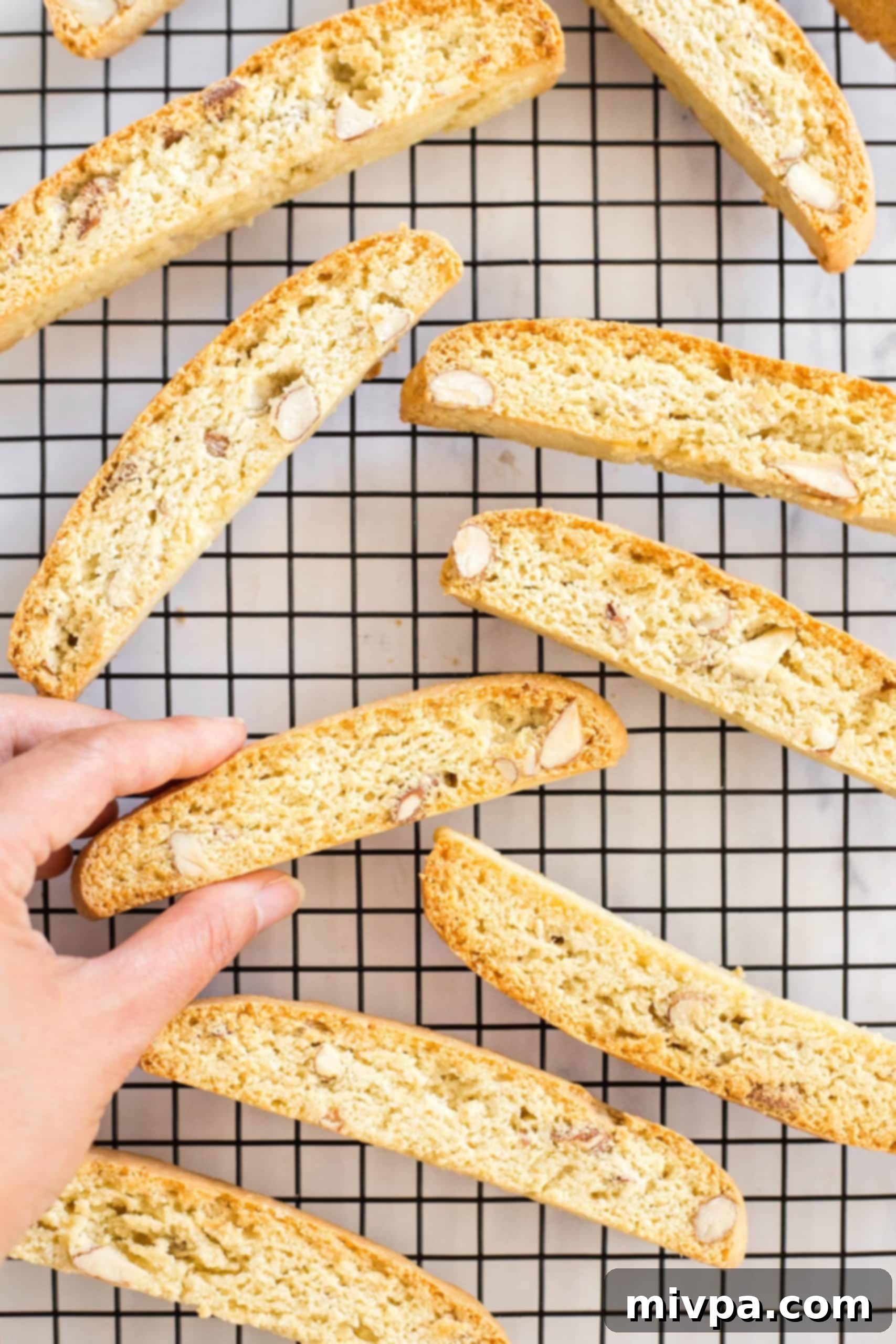 A hand reaching to pick up a perfectly cooled gluten-free almond biscotti from a wire rack, highlighting its readiness for enjoyment.