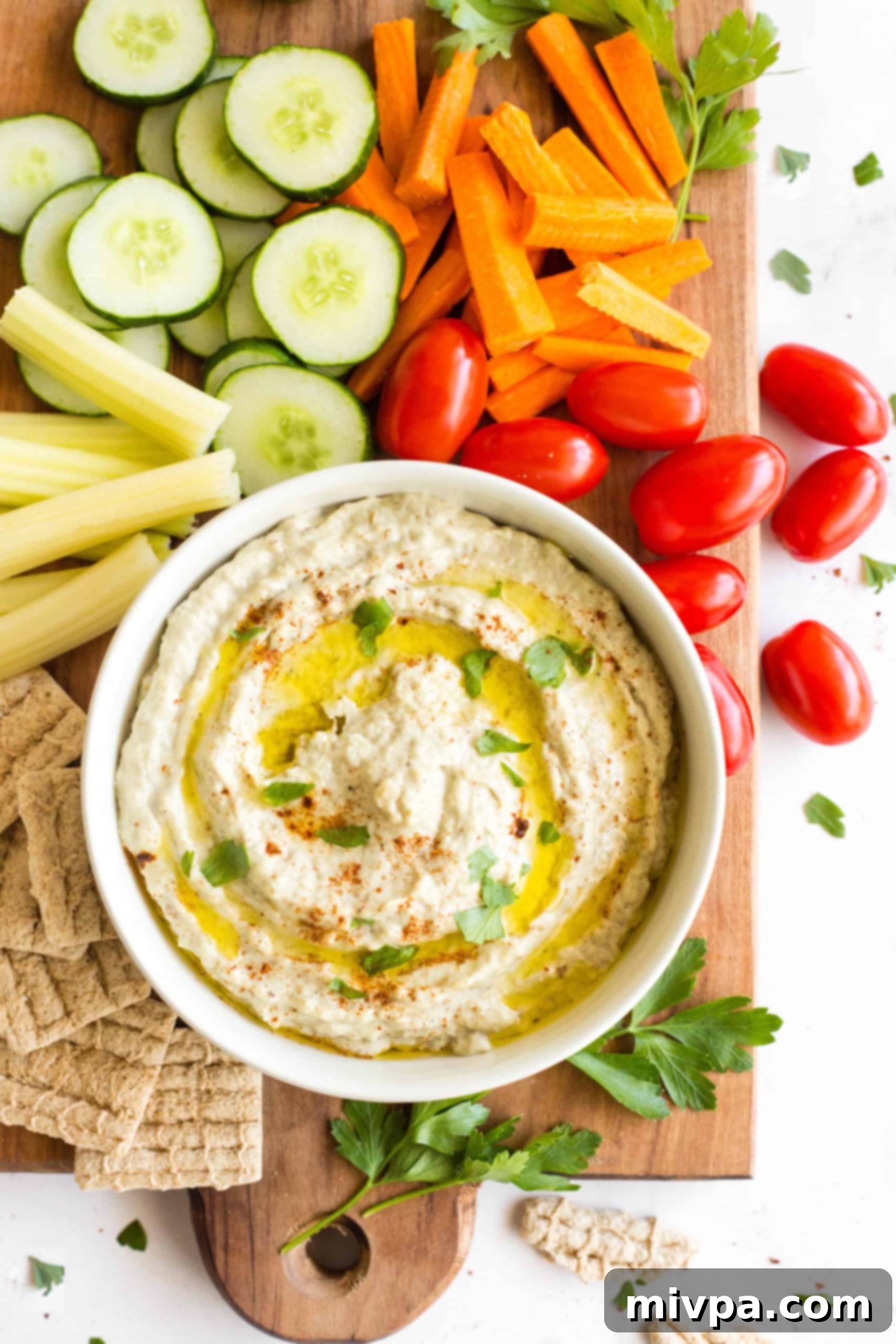 A wooden board with raw vegetable crudités, crackers, and eggplant dip.