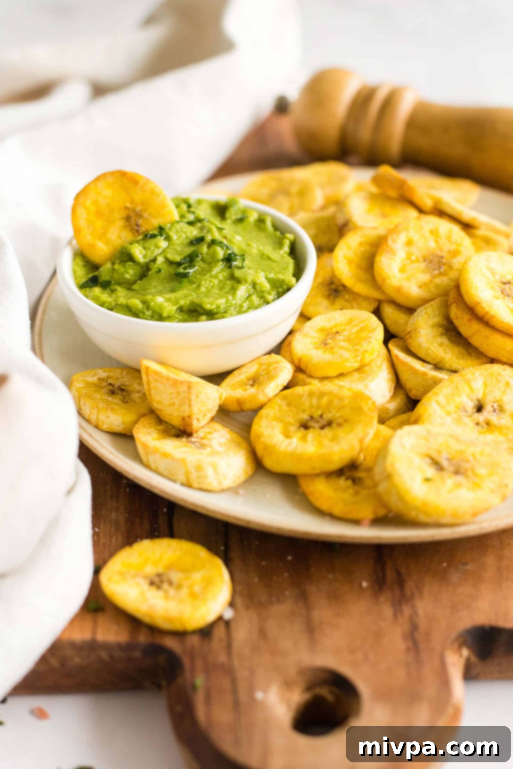 A plantain chip in a small bowl of guacamole alongside a plate of air fryer plantains.