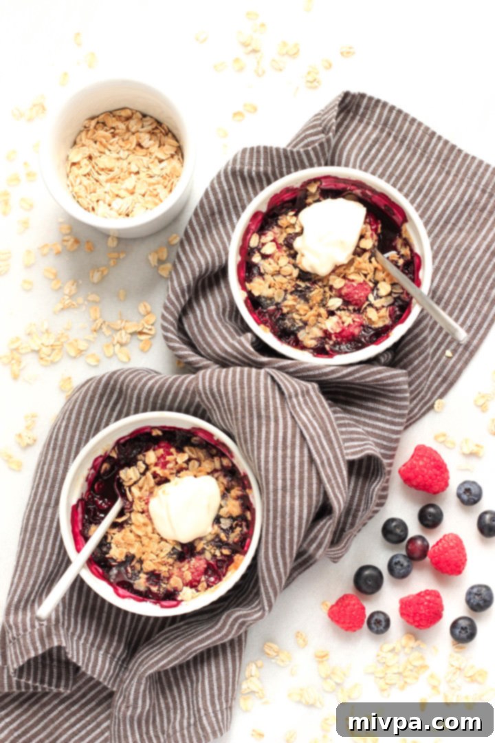 Gluten-free Berry Crisp (Dairy-Free, Vegan) Two ramekins of gluten-free berry crisp, generously topped with dairy-free ice cream, showcased on a wooden surface.