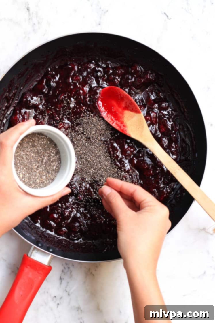 Mixed Berry Chia Seed Jam (Gluten-Free, Vegan) Sprinkling whole chia seeds over the cooked fruit mixture in a skillet, preparing to mix them in.