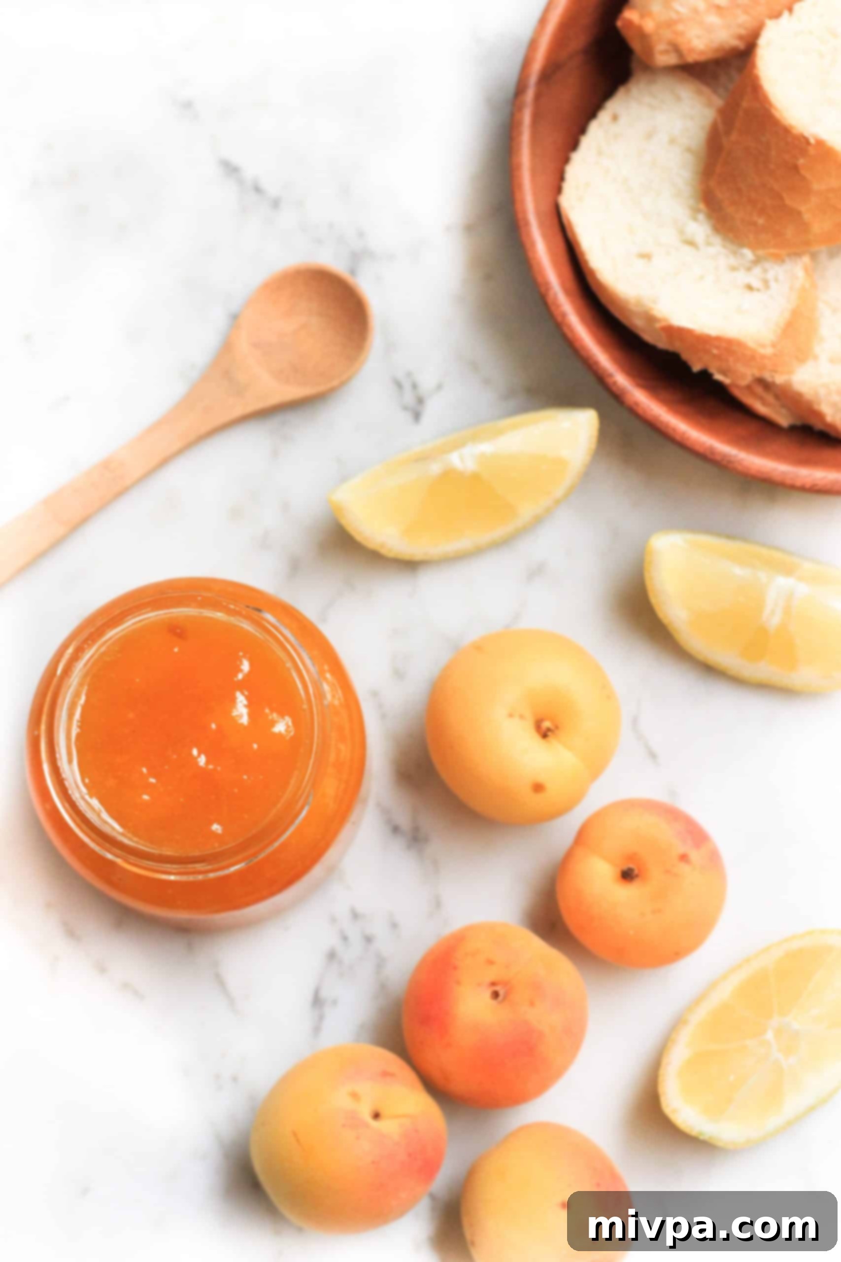An open jar of apricot jelly, apricots, lemons, and bread on a marble board.