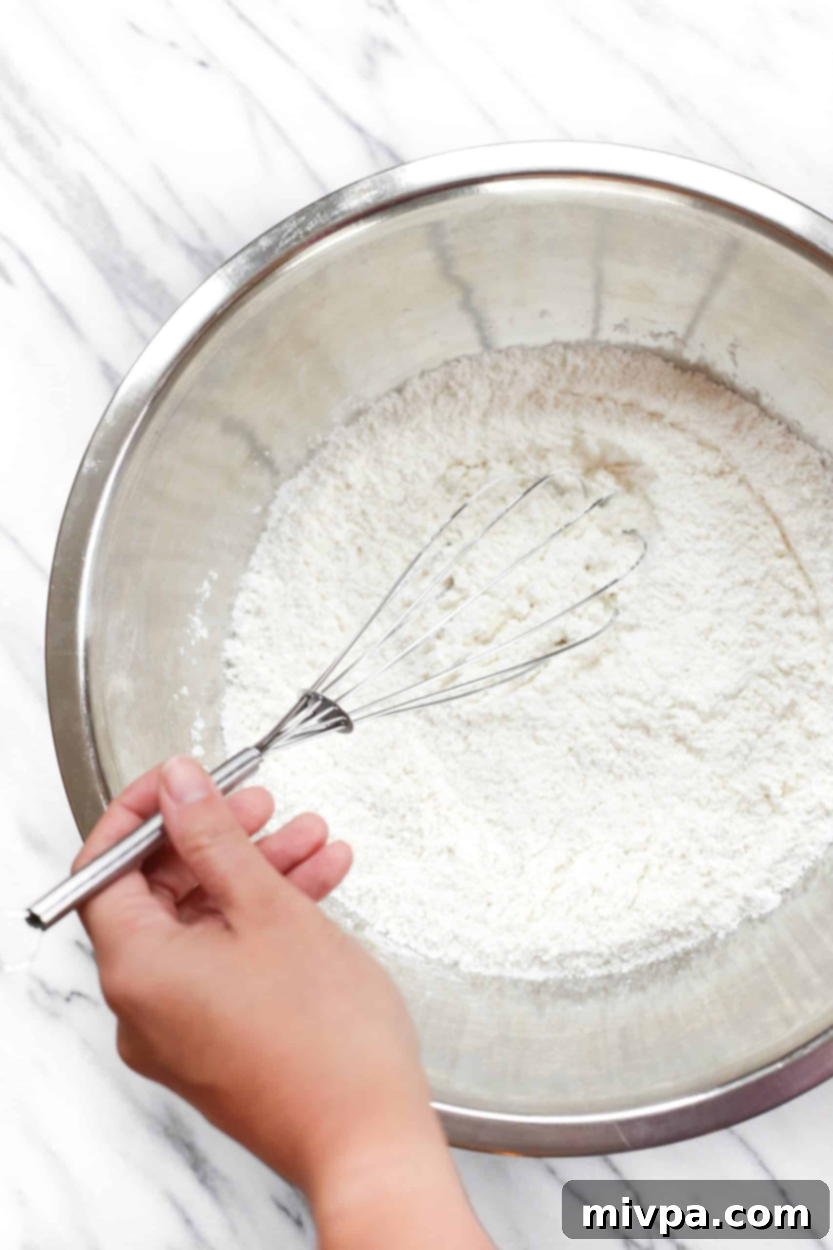 A hand whisking gluten-free flour, baking powder, and salt in a metal bowl.