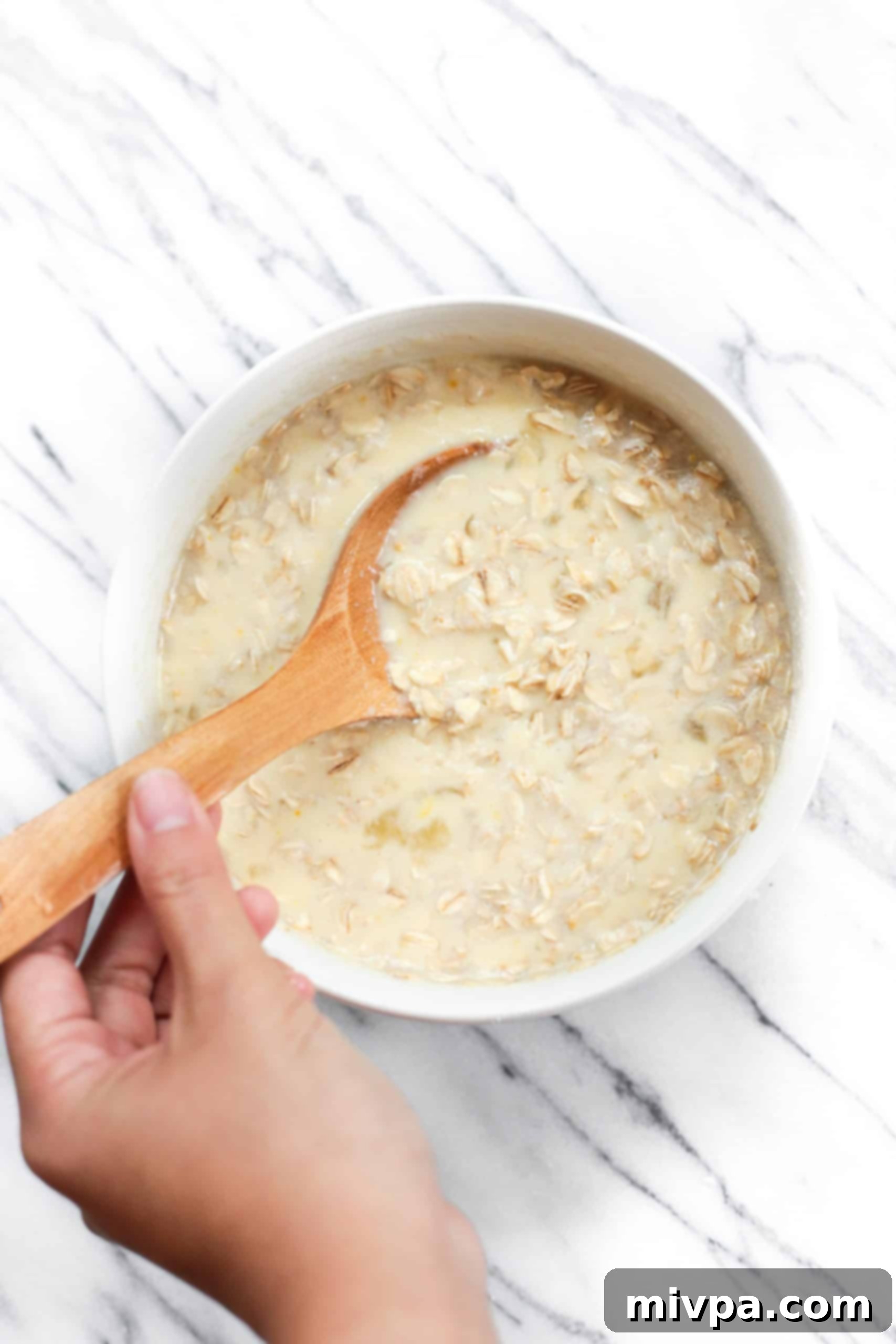 A hand mixing wet ingredients with a wooden spoon in a white bowl, showing egg, yogurt, oil, honey, and oats.