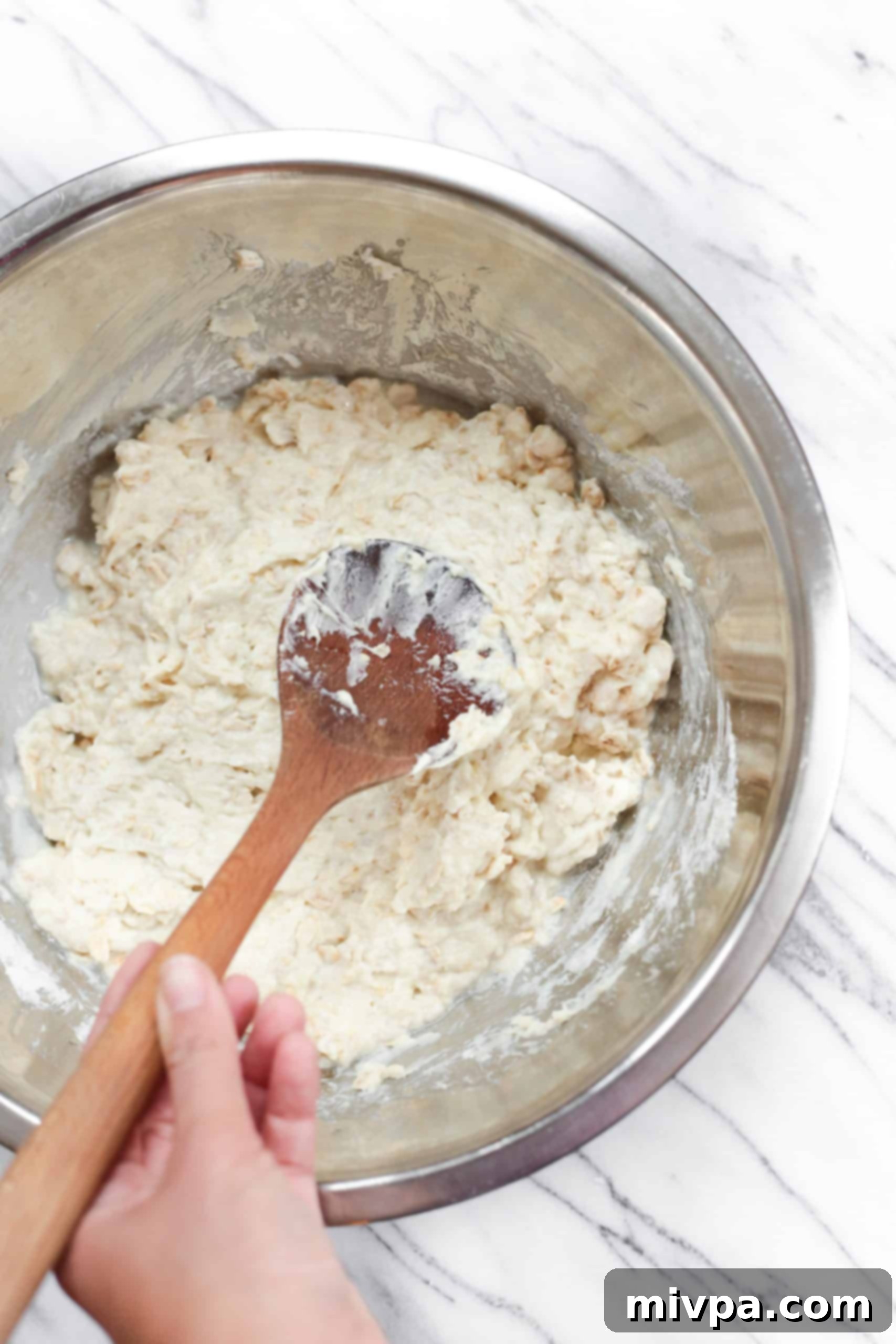 A hand mixing thick batter with a wooden spoon in a metal bowl, showing the combined wet and dry ingredients.