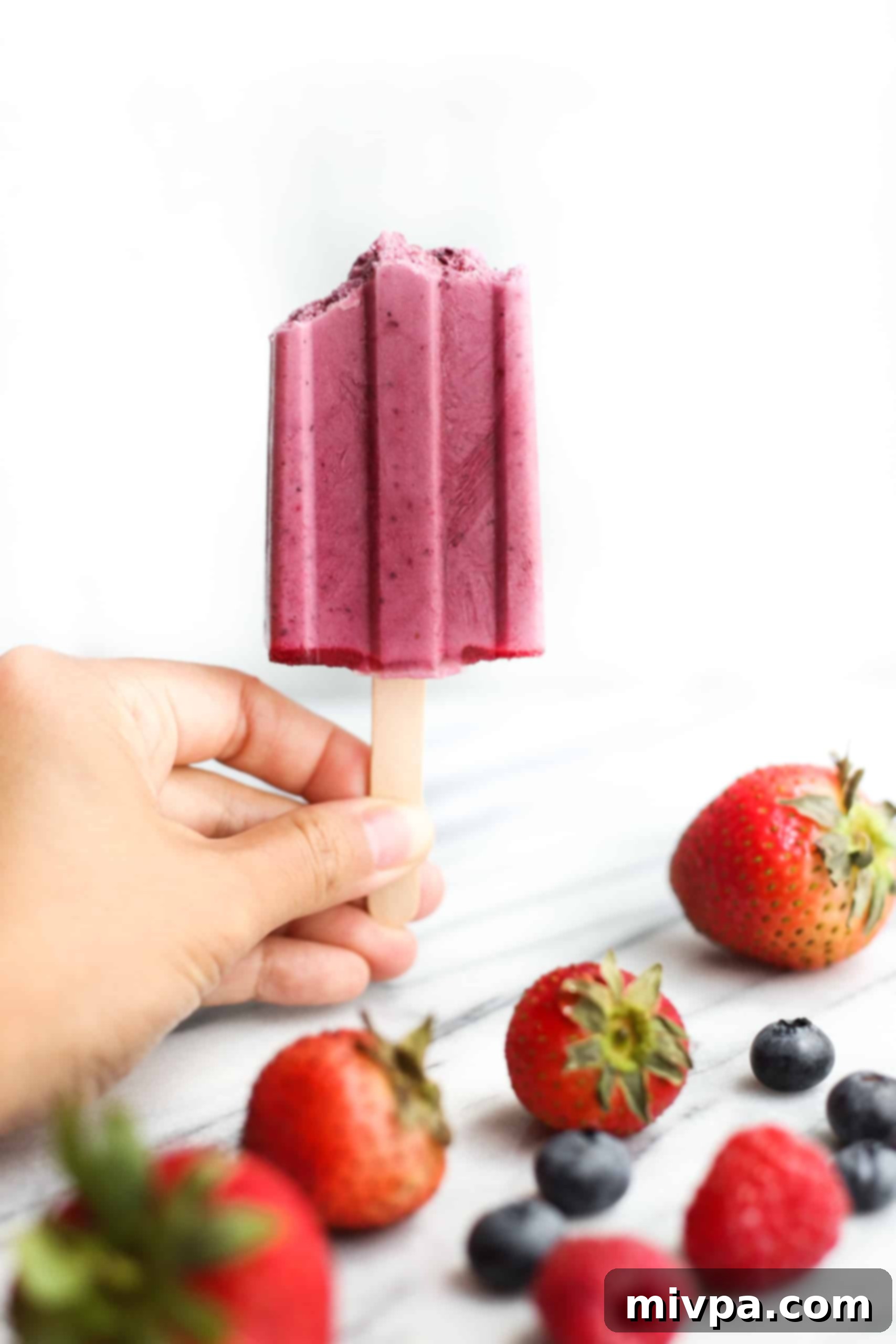 A hand holding up a homemade berry yogurt popsicle with fresh berries visible within the frozen treat.