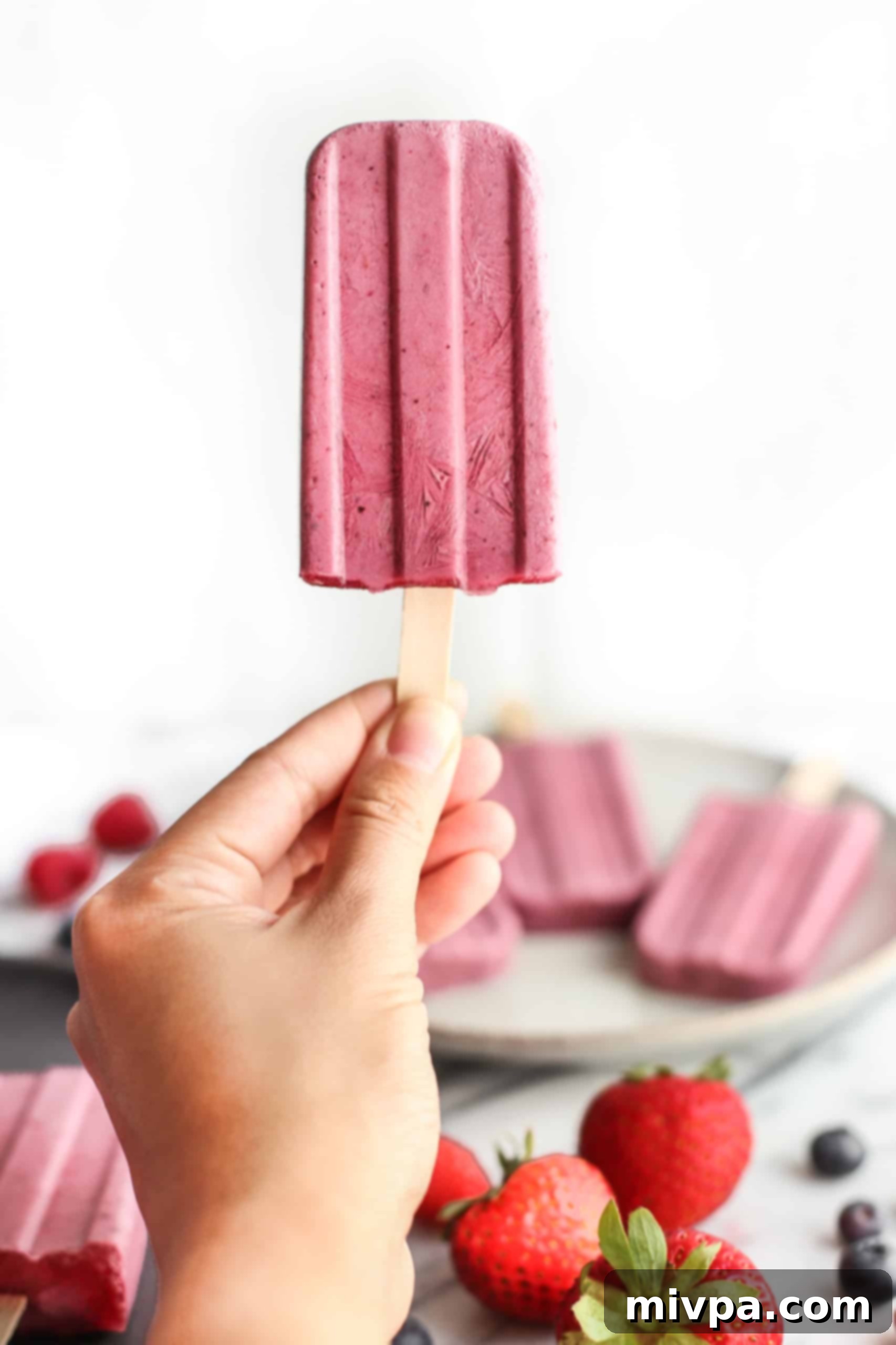 A hand holding up a single homemade berry yogurt popsicle, showing its perfect shape and inviting texture.