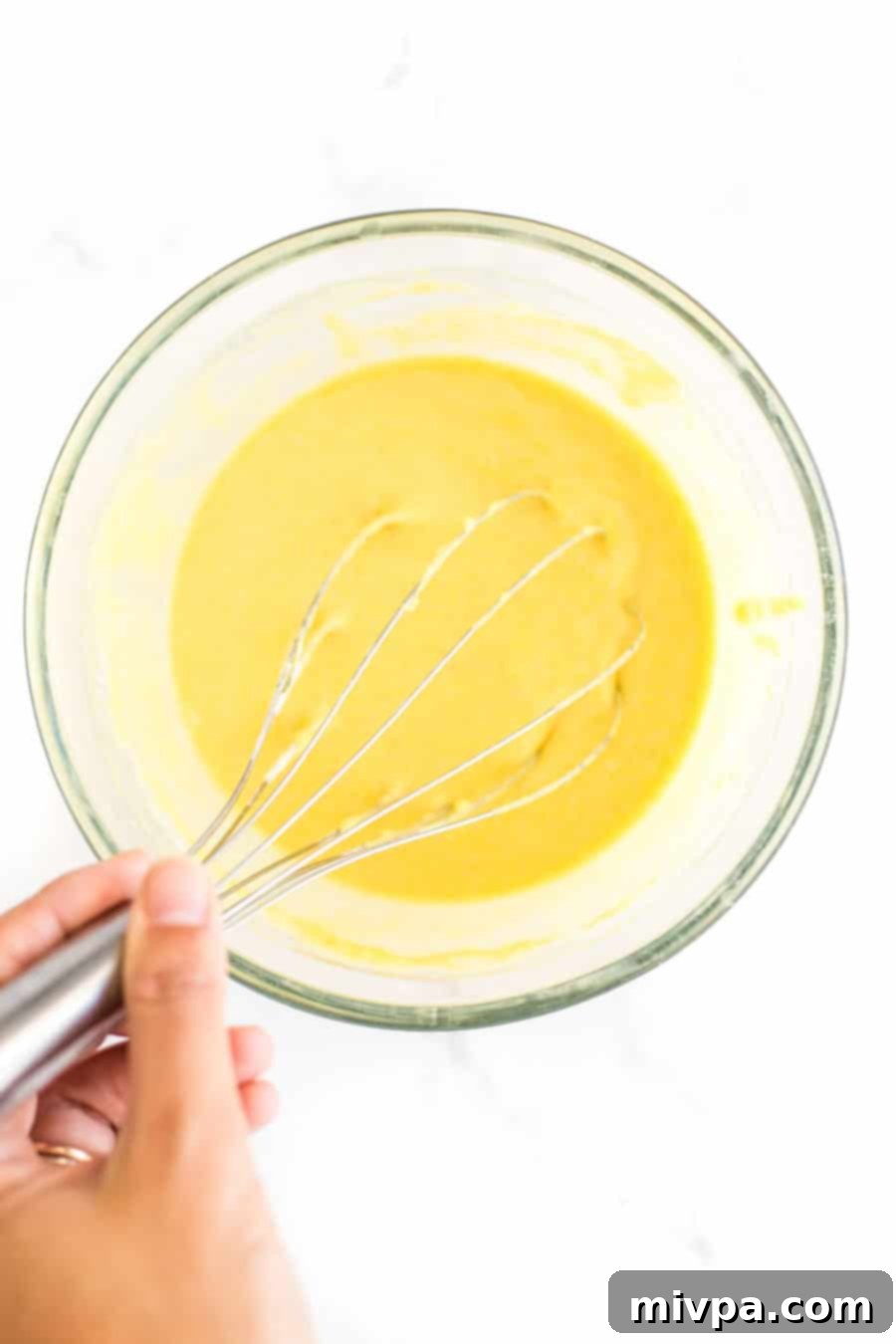 Whisking golden corn flour pancake batter in a clear glass bowl, showcasing its smooth consistency.