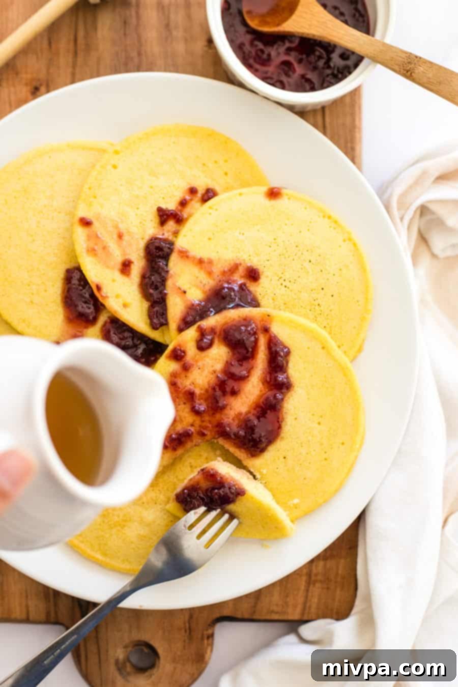 A close-up of maple syrup being poured over a stack of corn flour pancakes, glistening deliciously.