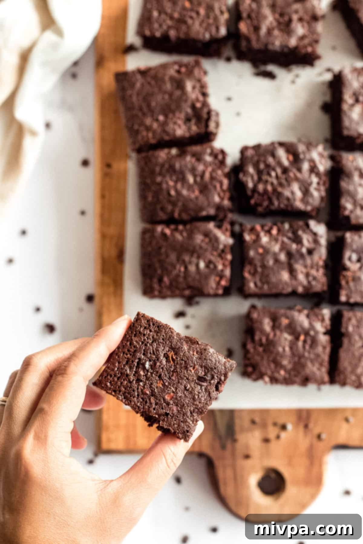 A hand picking up a single fudgy gluten-free dairy-free brownie from a rustic wooden board, emphasizing its irresistible appeal and deliciousness.