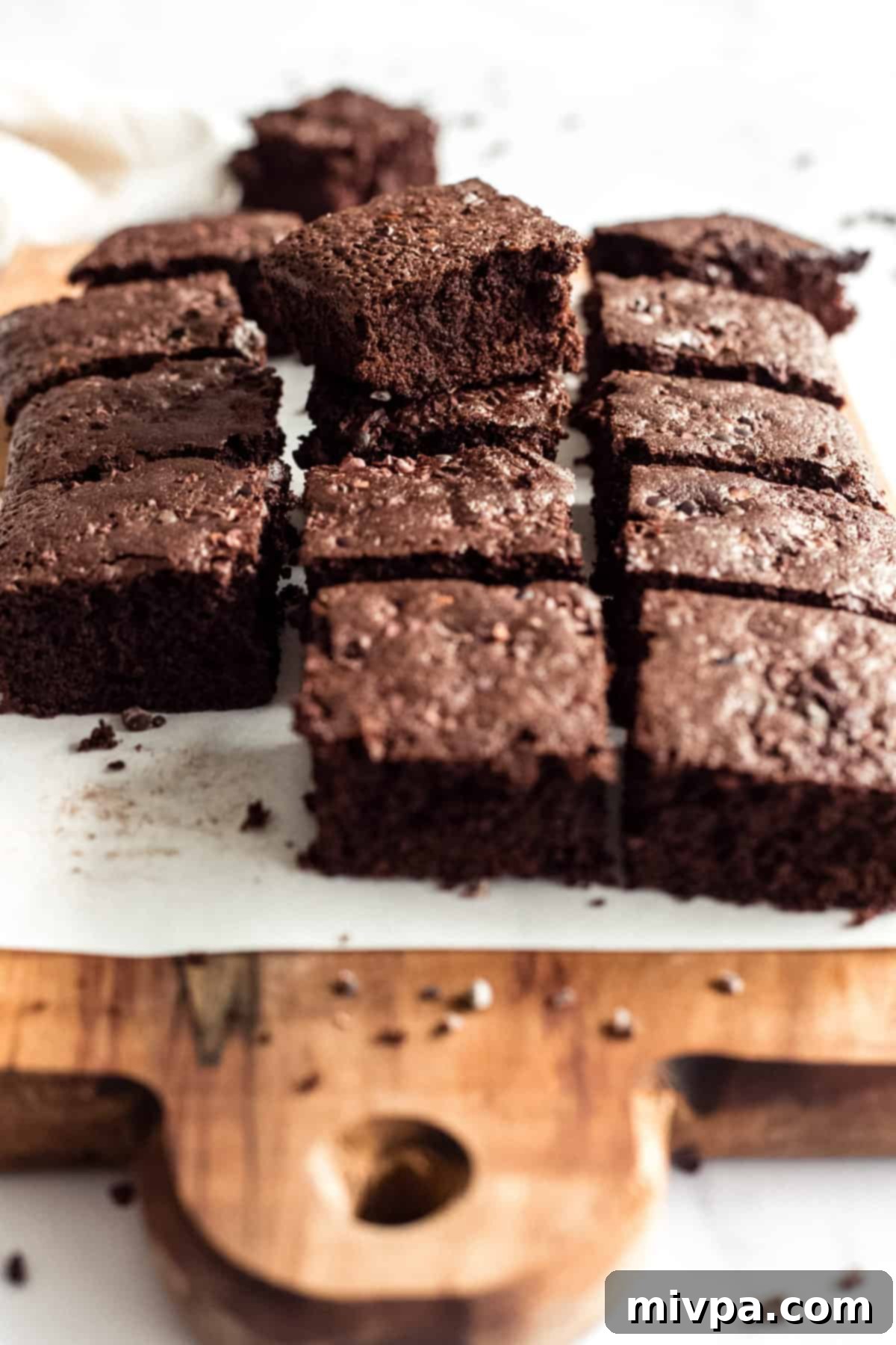 Multiple fudgy gluten-free dairy-free brownies arranged on a parchment-lined wooden cutting board, showcasing their rich texture and deep color.