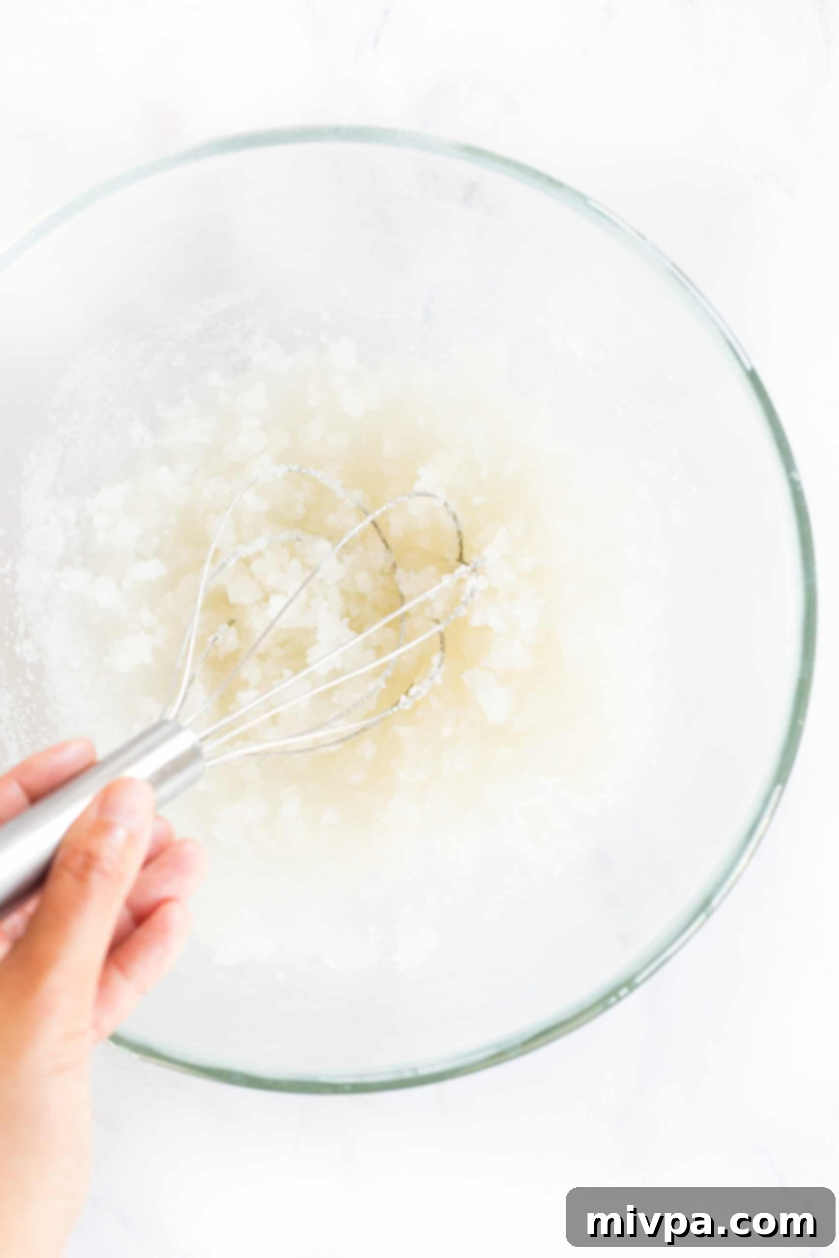 A whisk blending granulated sugar into warm sunflower oil in a mixing bowl, creating a smooth, glossy base for the brownie batter.