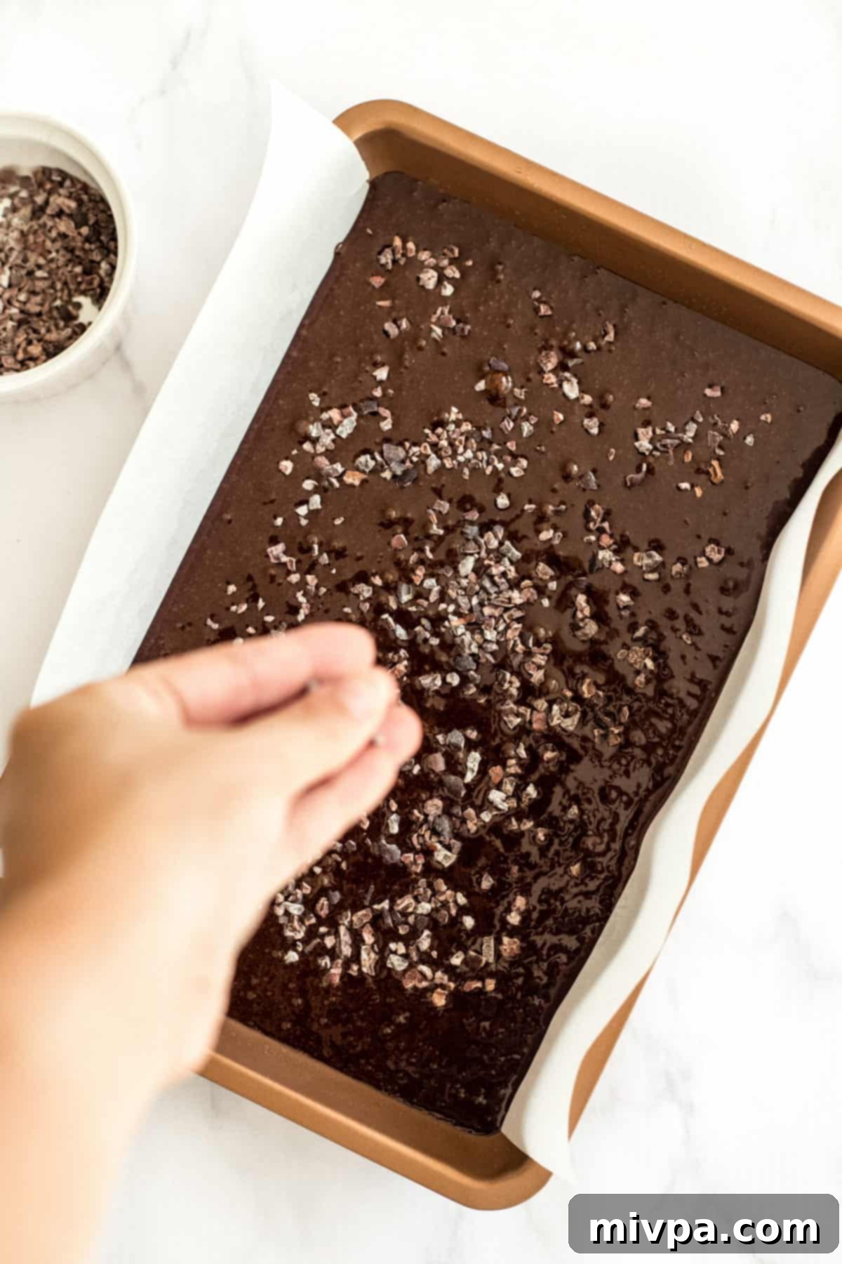 A baking pan filled with rich gluten-free dairy-free brownie batter, topped with an even sprinkle of cacao nibs, ready for the oven.