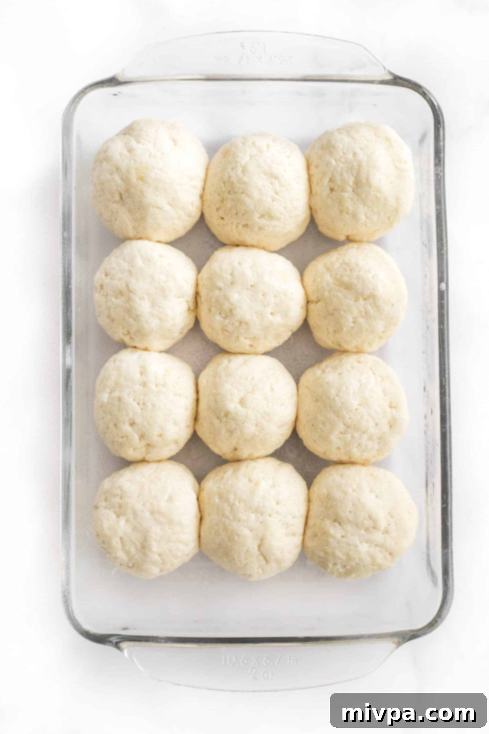 Close-up of small balls of gluten-free bread dough visibly rising in a glass baking pan, covered lightly.