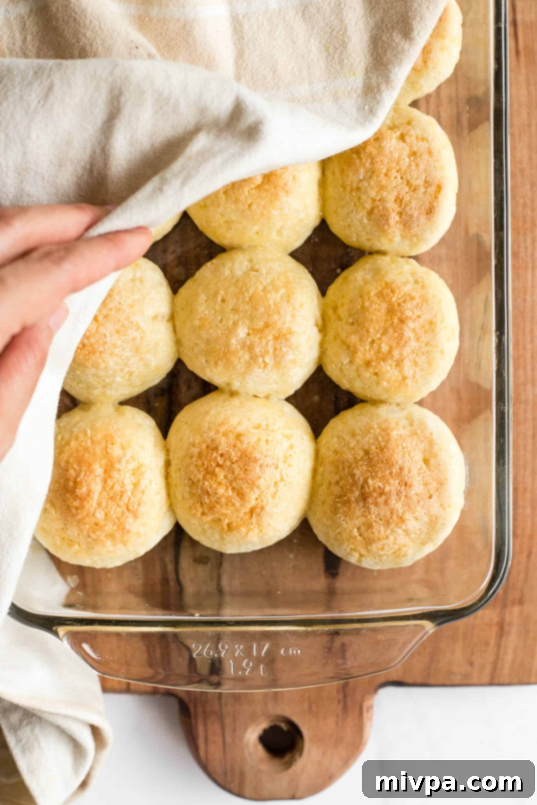 A hand removing a kitchen towel, revealing a pan full of perfectly risen and baked gluten-free dinner rolls.
