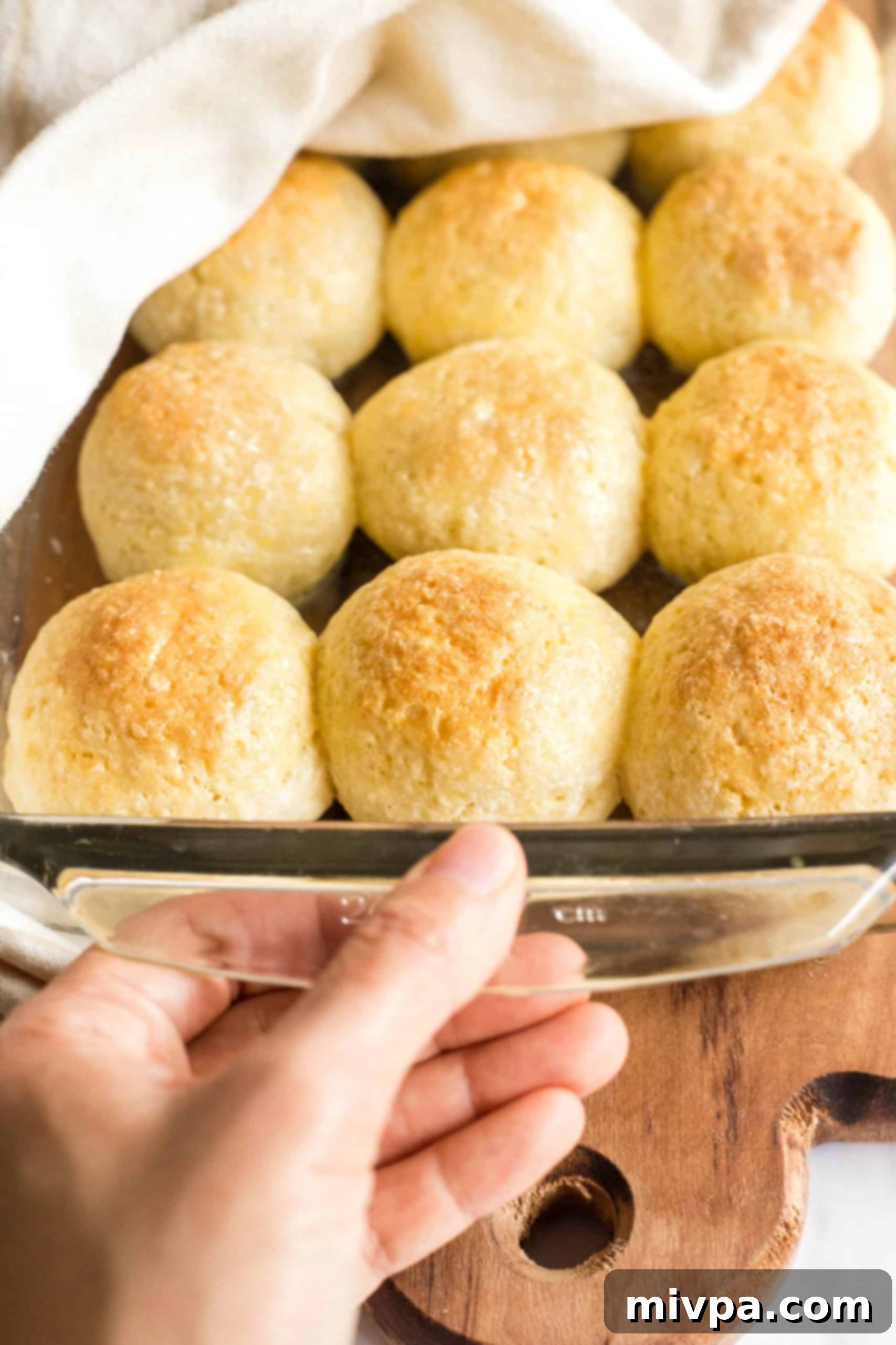 A hand holding a glass baking pan filled with a batch of freshly baked, soft gluten-free yeast rolls.