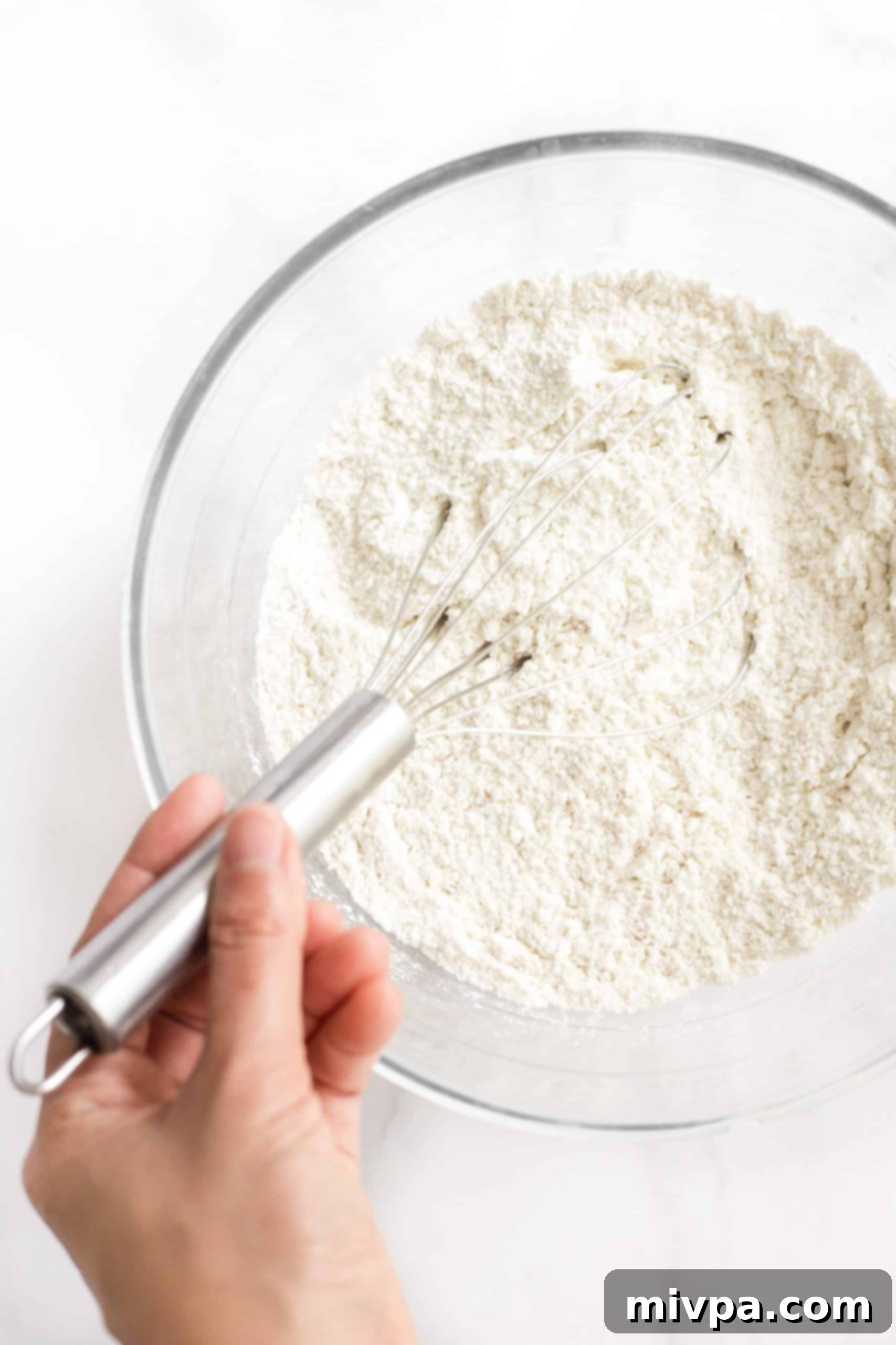 A hand whisking gluten-free flour, xanthan gum, salt, baking powder, sugar, and instant dry yeast in a clear glass mixing bowl.