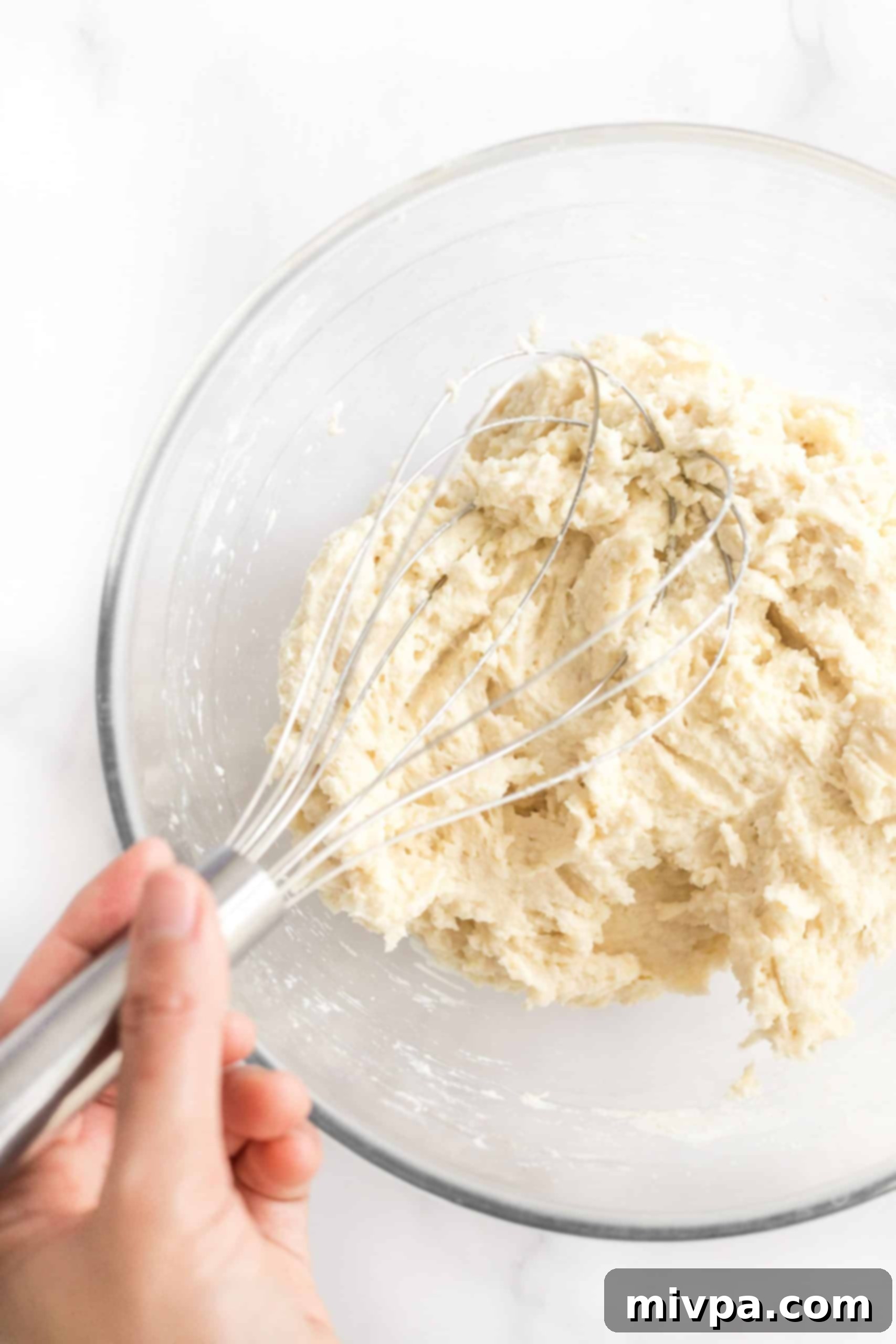 Mixing the thick gluten-free bread dough with a spoon in a clear glass mixing bowl after adding wet ingredients.