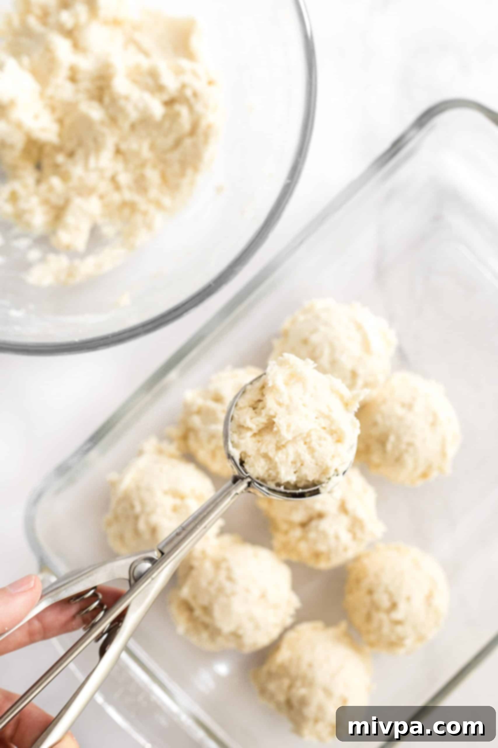 A hand scooping gluten-free bread dough with an ice cream scoop into a glass baking pan.
