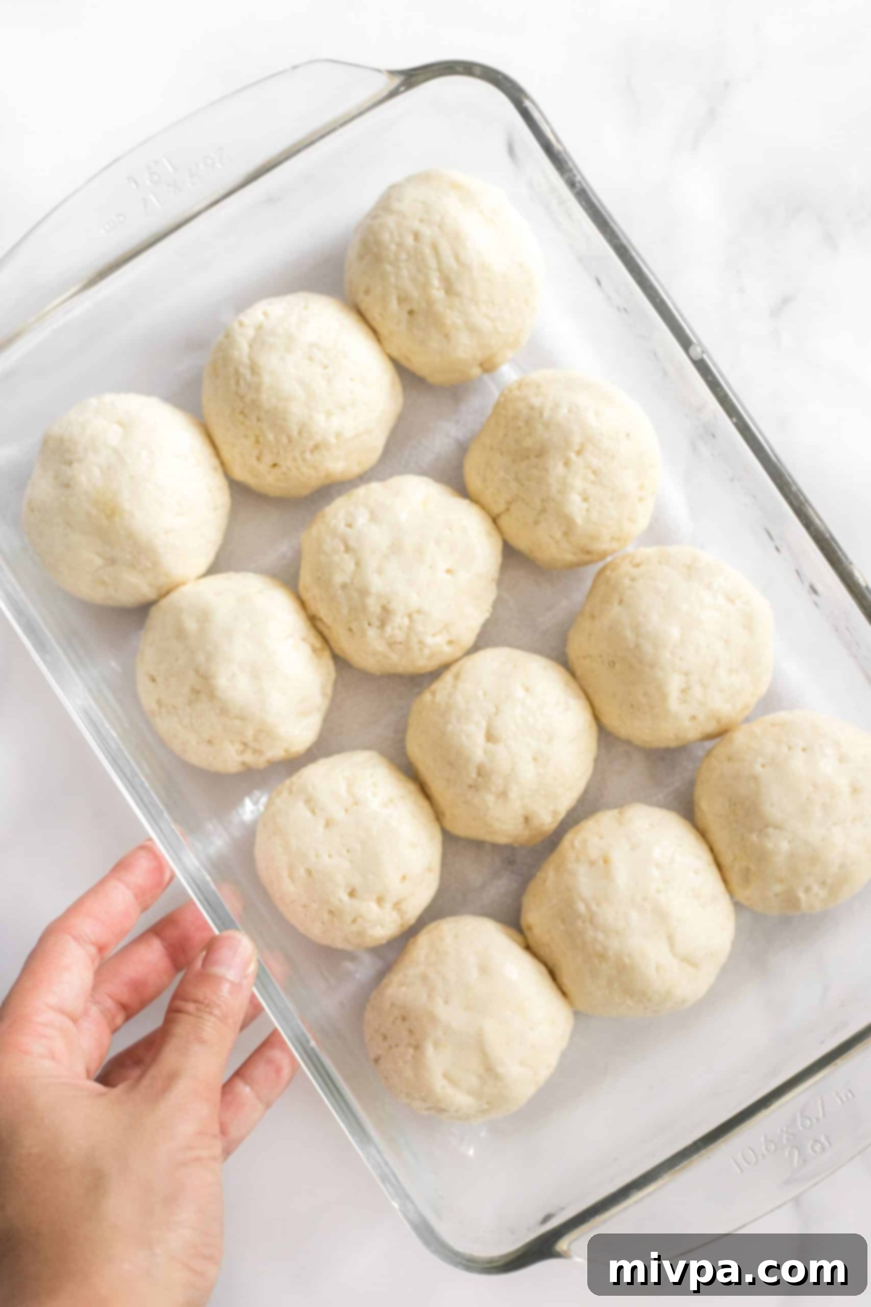 A hand holding a glass baking pan filled with neatly shaped balls of gluten-free bread dough, ready for rising.