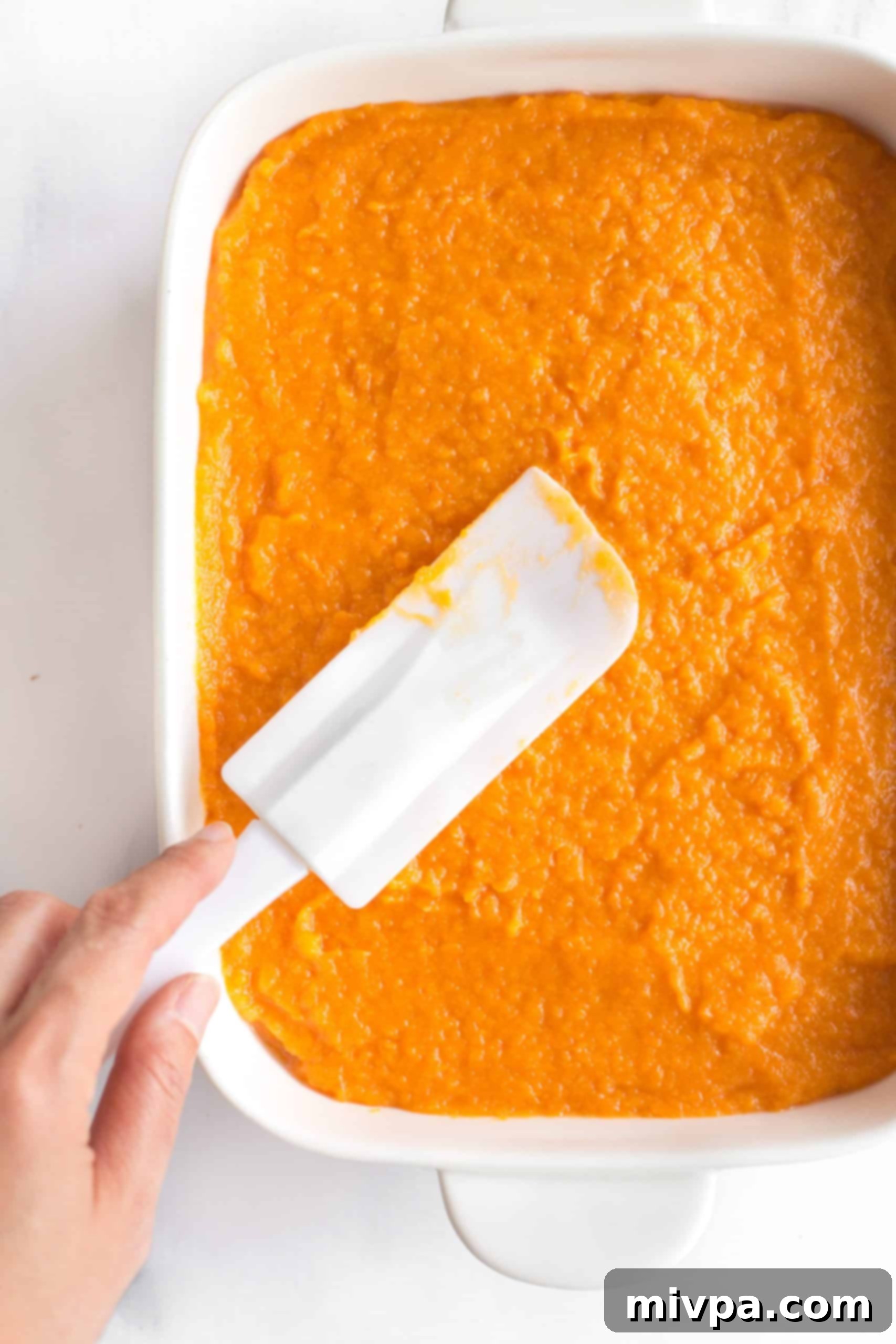 Sweet Potato Casserole Recipe (Gluten-Free, Dairy-Free) Smoothing out sweet potato filling with a spatula in a white casserole dish.