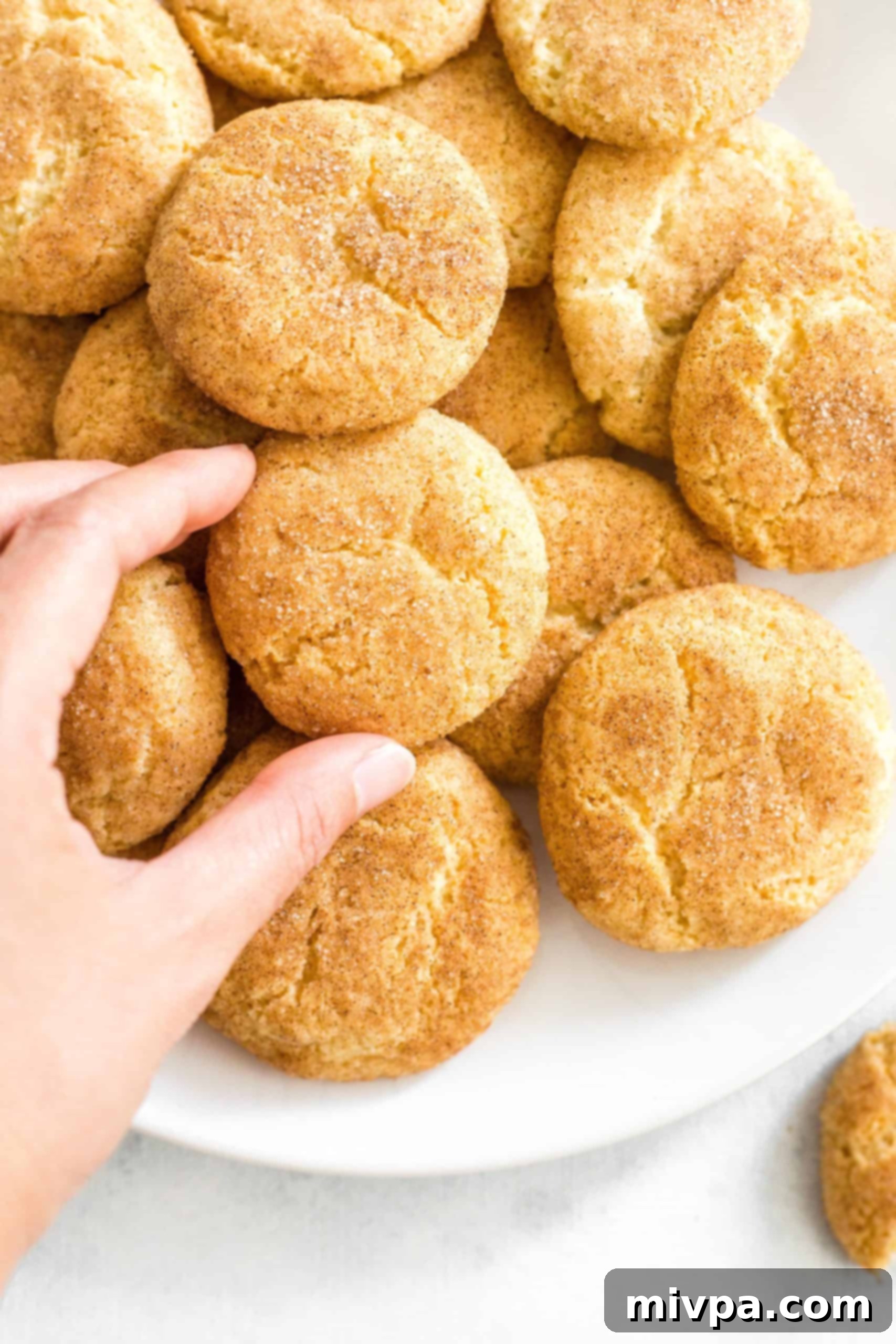 A hand reaching for a freshly baked gluten-free and dairy-free snickerdoodle cookie from a plate piled high with more delicious cookies.
