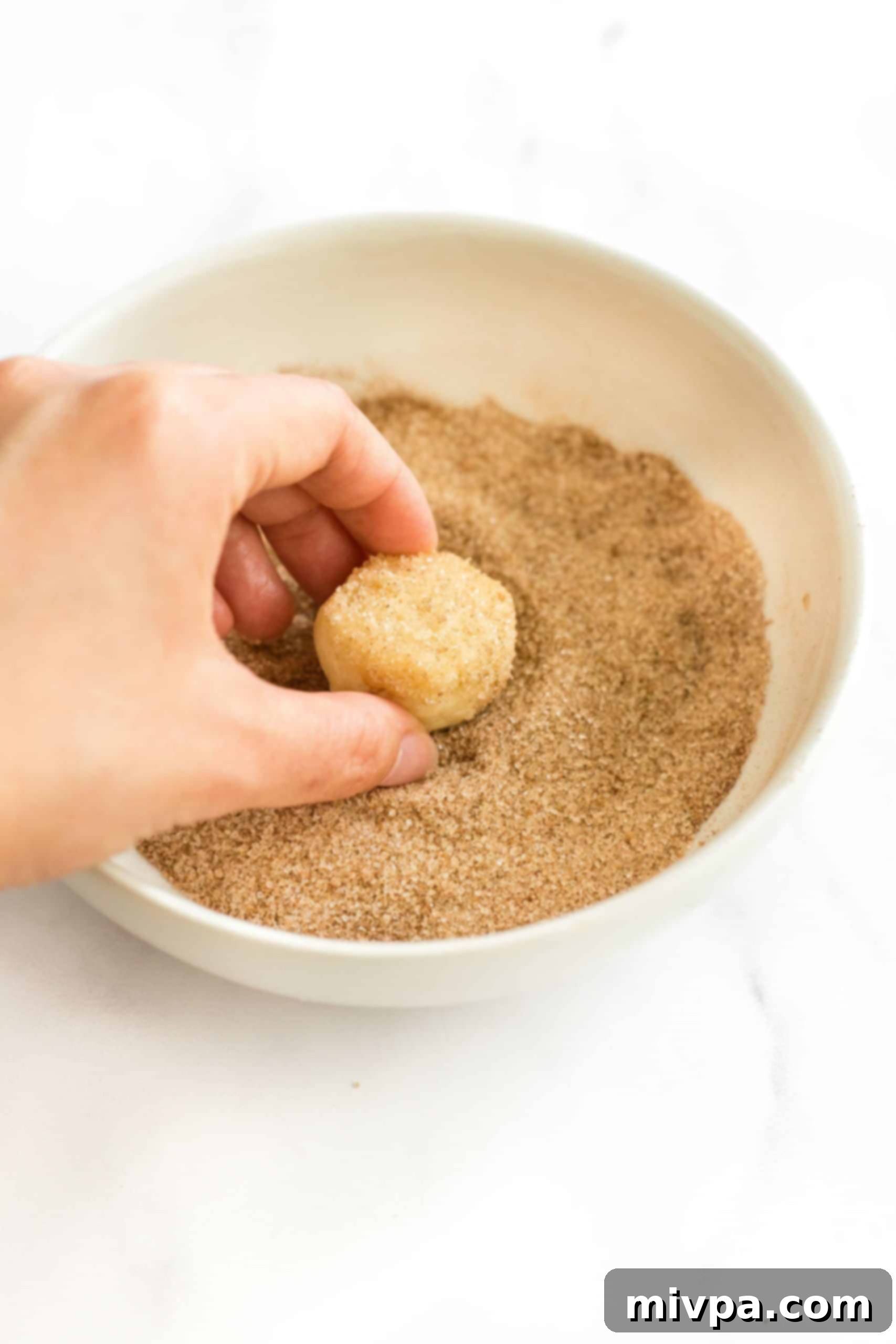A hand rolling a gluten-free snickerdoodle cookie dough ball in a shallow bowl of cinnamon sugar mixture, ensuring a full coating.