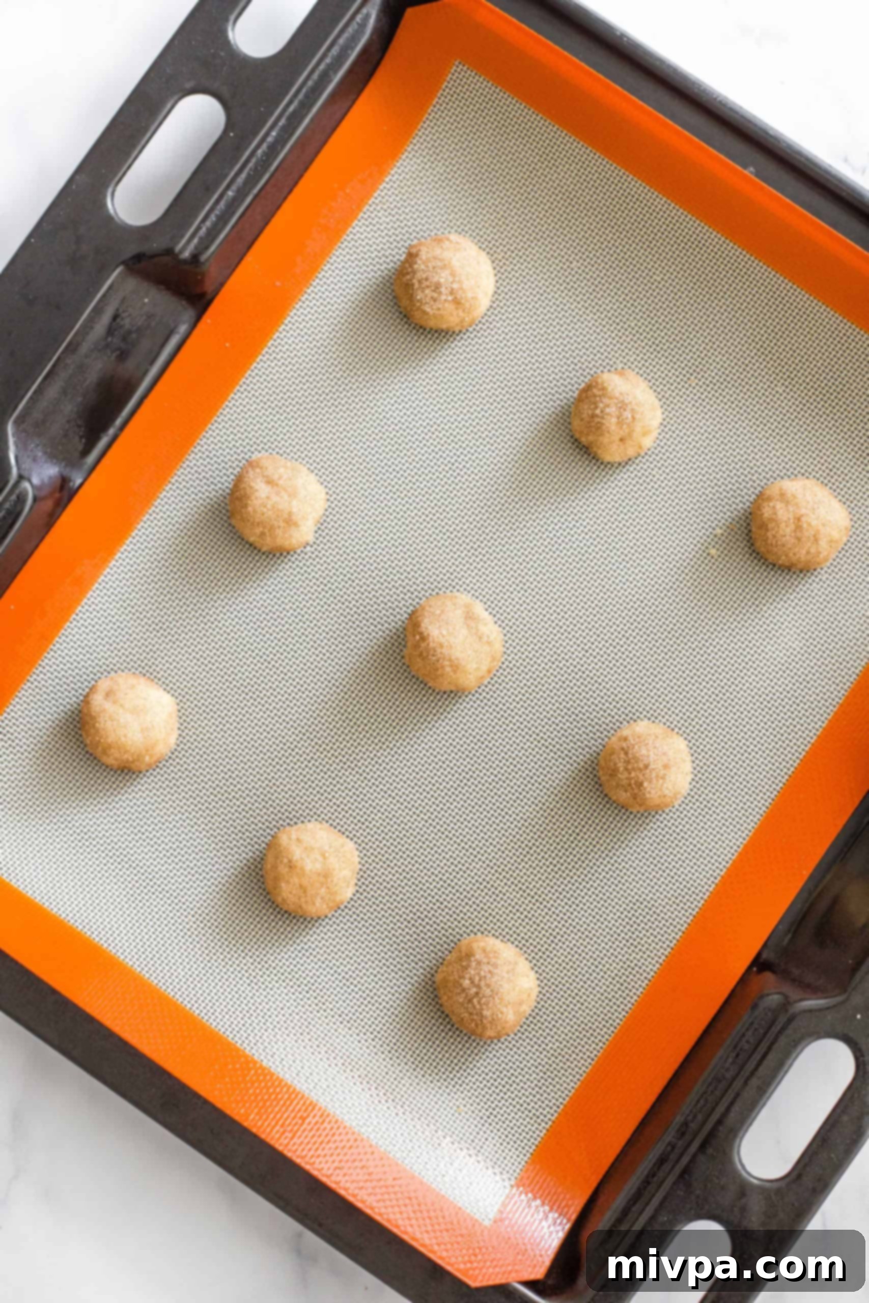 Gluten-free snickerdoodle cookie dough balls, coated in cinnamon sugar, arranged on a Silpat-lined baking sheet, ready for baking.