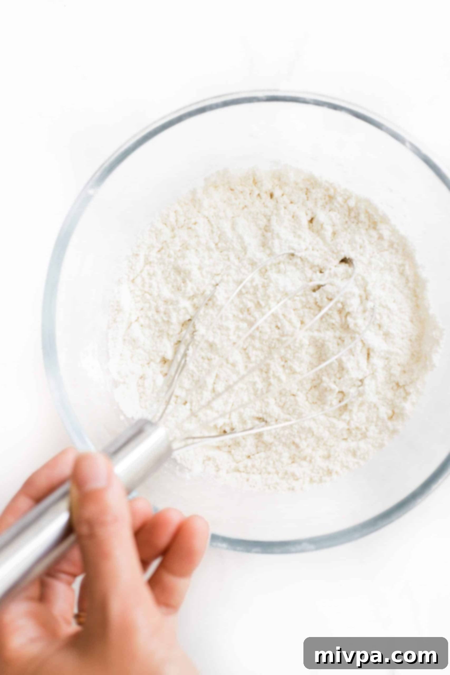 A hand whisking gluten-free flour, cream of tartar, baking soda, and salt in a clear glass mixing bowl for snickerdoodle cookies.