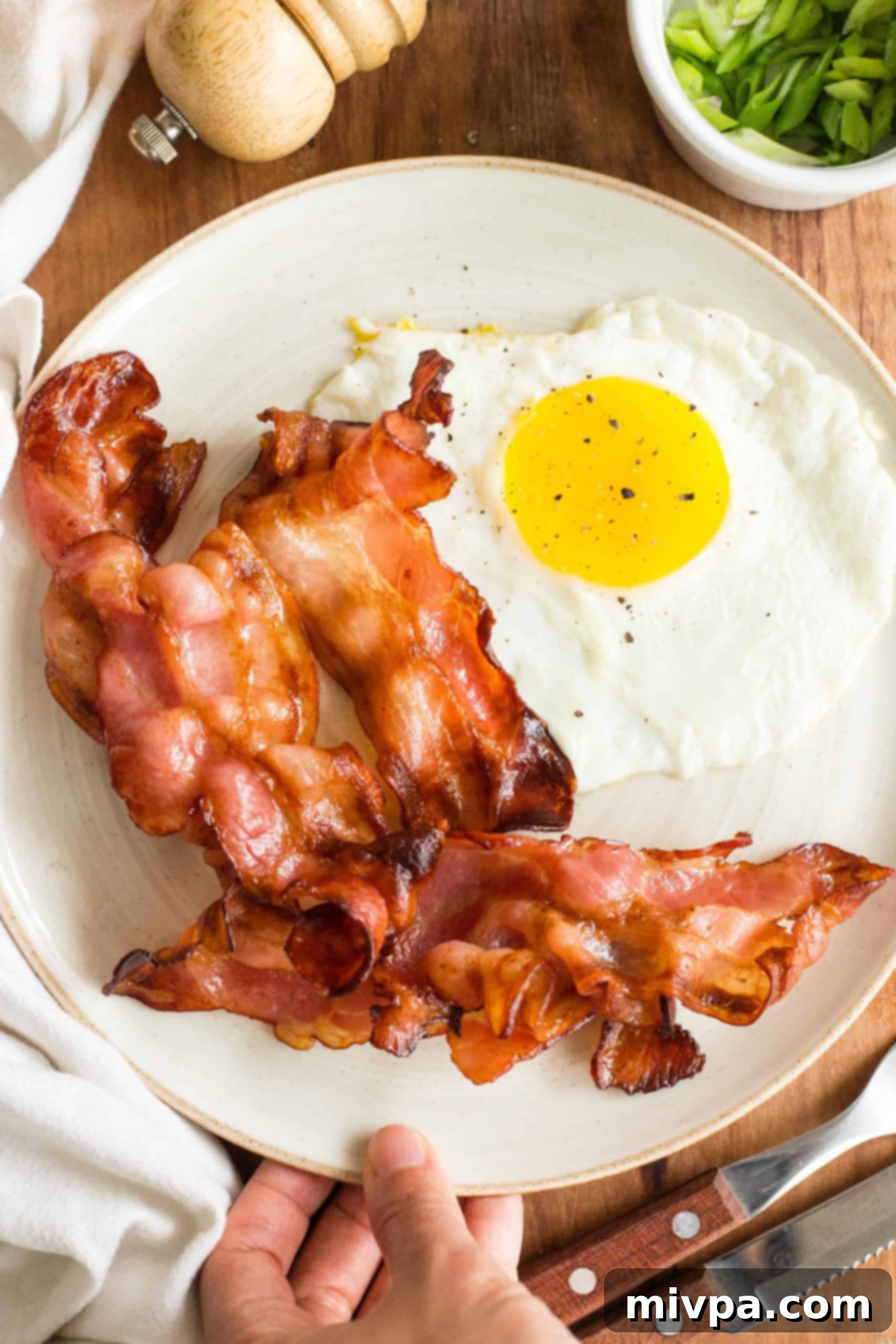Hand holding a plate with air fried bacon and eggs, showcasing crispy bacon and a perfectly cooked egg.