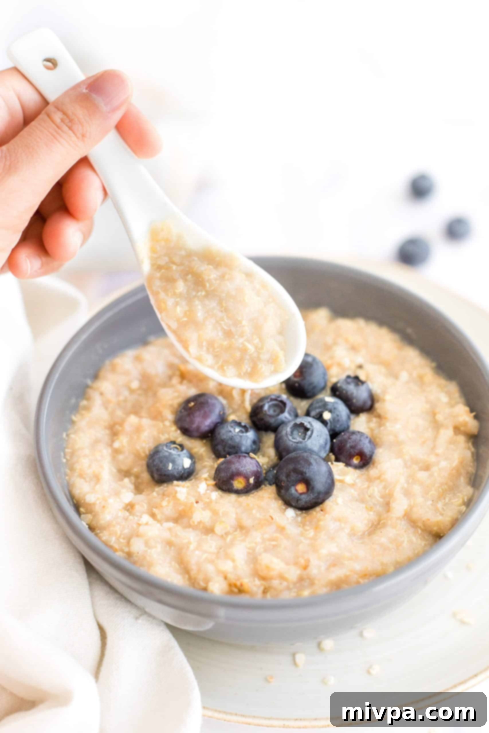 Holding up a spoonful of creamy quinoa porridge from a grey bowl, garnished with fresh blueberries and a mint leaf.