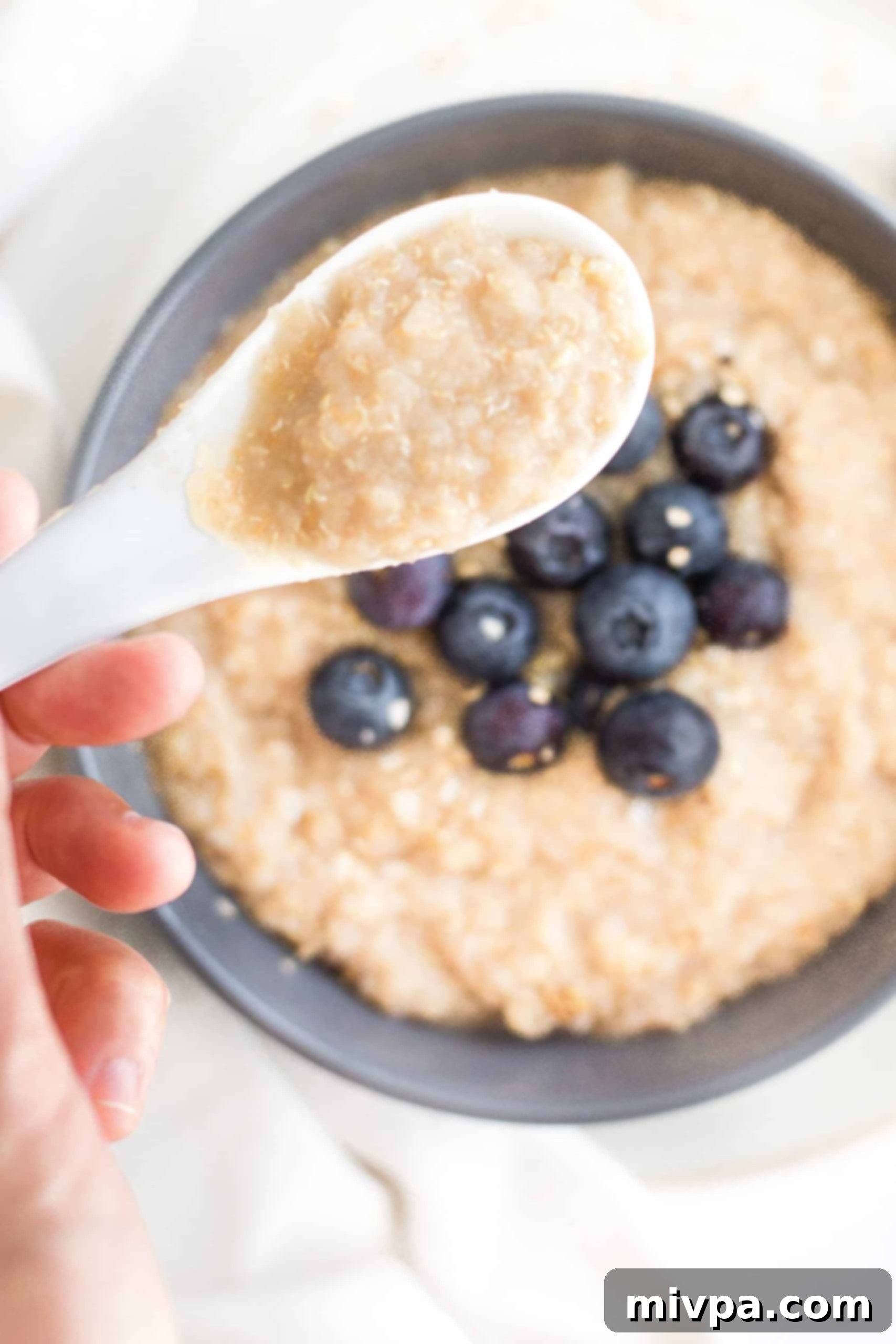 A hand holding a spoonful of creamy quinoa porridge, garnished with blueberries, showcasing its inviting texture and delicious appeal.