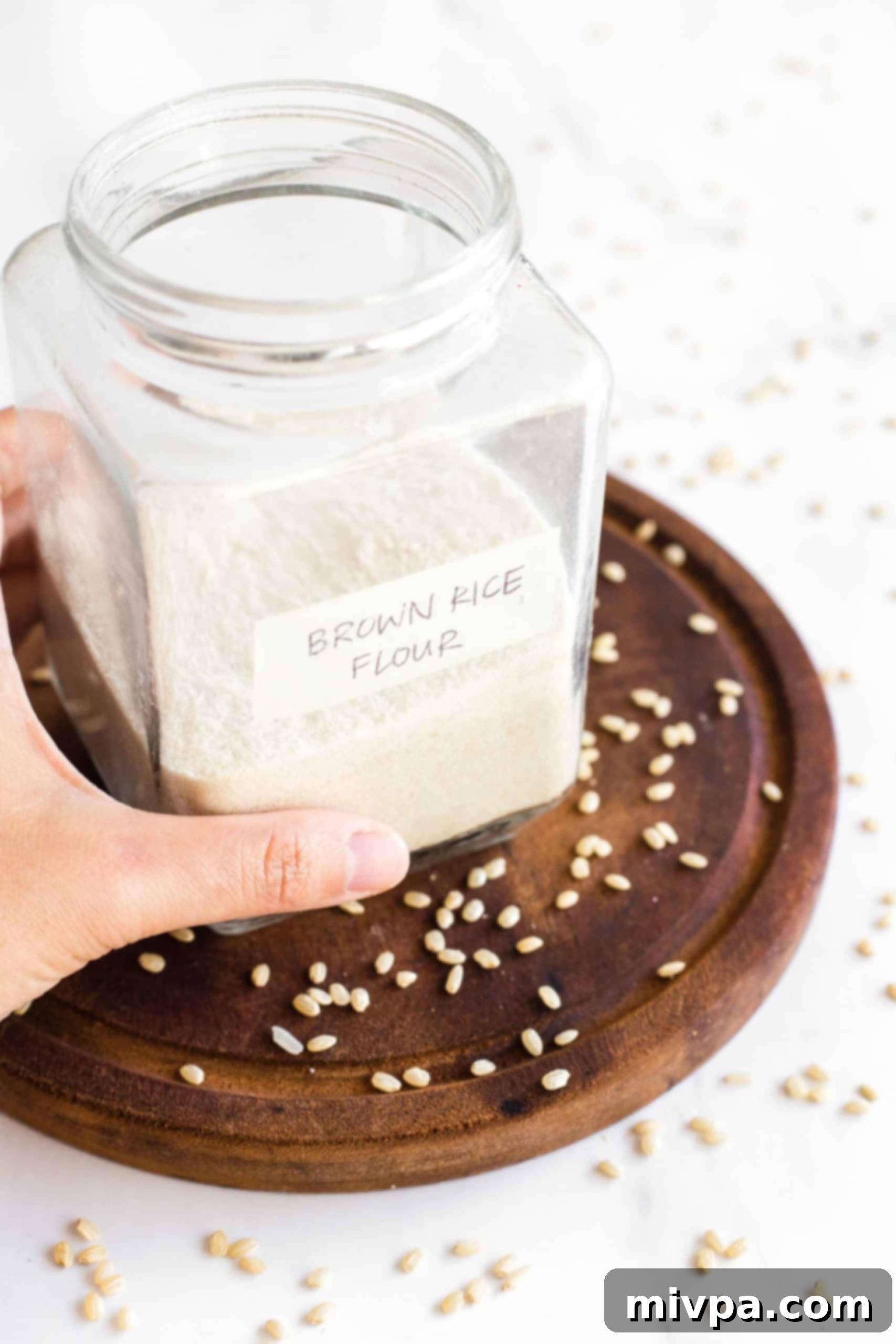 A half-filled jar of homemade brown rice flour sits on a wooden board, surrounded by loose brown rice grains, highlighting the transformation.