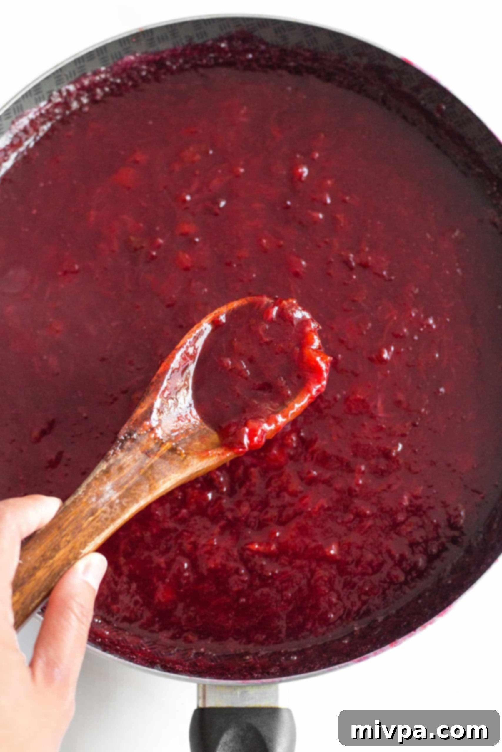 Plum Jelly Without Pectin (Gluten-Free, Vegan, Pectin-Free) A wooden spoon holding a spoonful of thick plum jam over a simmering skillet, demonstrating the perfect jelling point.