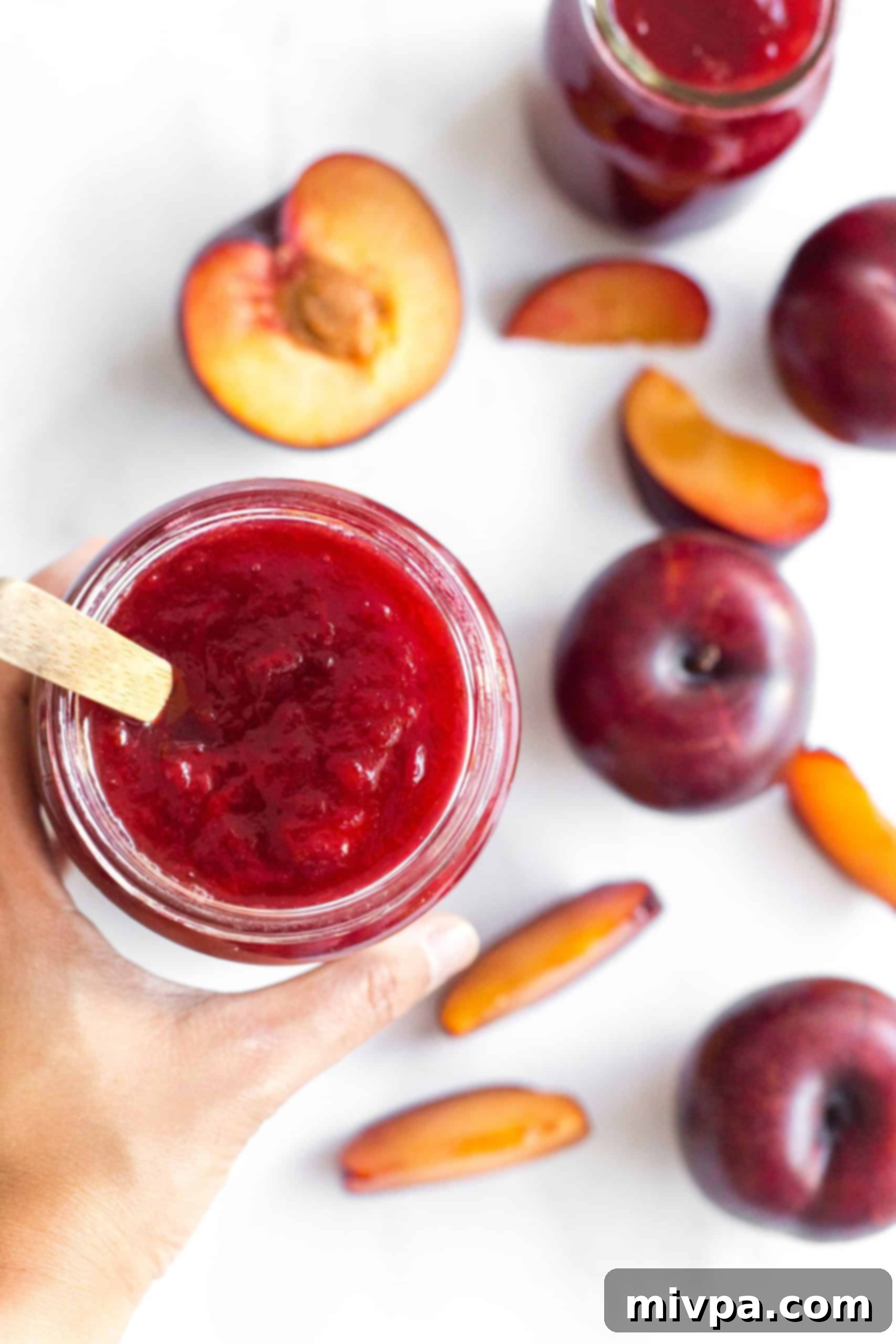 Easy Plum Jam Recipe (Gluten-Free, Vegan, Pectin-Free) A hand gently holding an open jar of glistening plum jelly, surrounded by fresh whole plums, symbolizing the pure essence of fruit captured in a jar.
