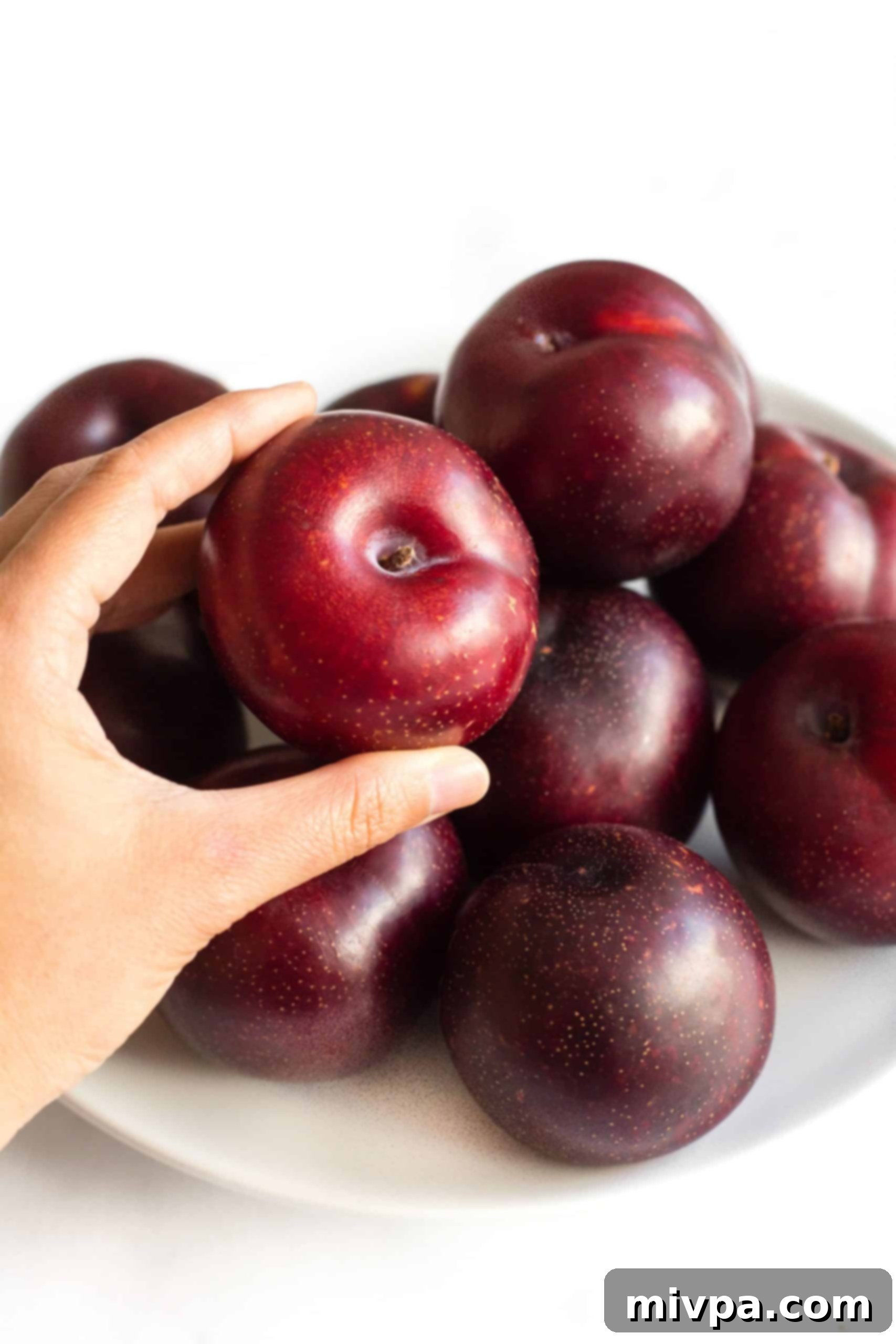 Easy Red Plum Jam Recipe (Gluten-Free, Vegan, Pectin-Free) A hand reaching for a fresh, ripe red plum from a rustic plate overflowing with plums, symbolizing the bounty of stone fruit season.