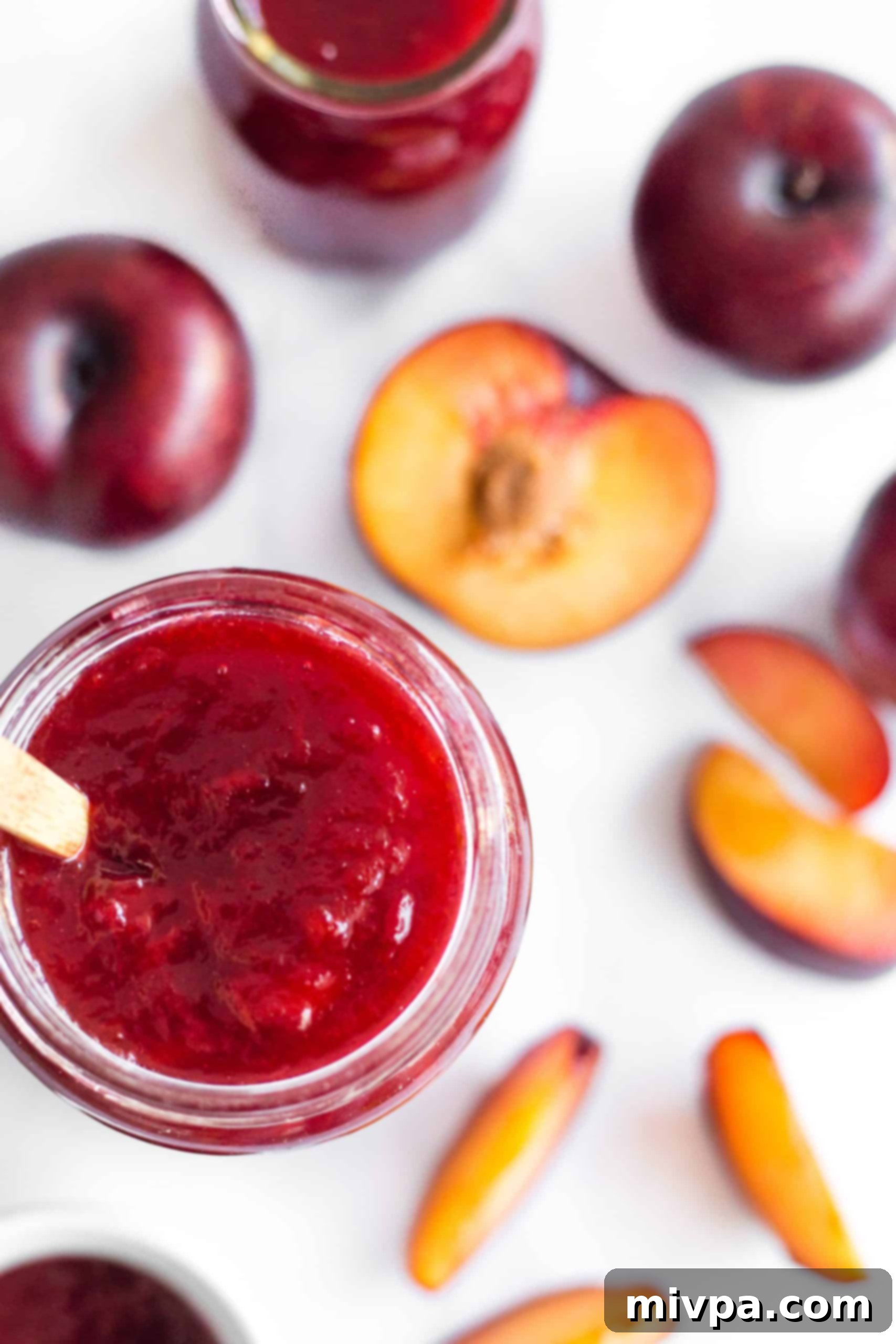 Easy Plum Jam Recipe (Gluten-Free, Vegan, Pectin-Free) An inviting open jar of homemade plum jelly, its deep red color shimmering, surrounded by fresh plum slices on a rustic surface.