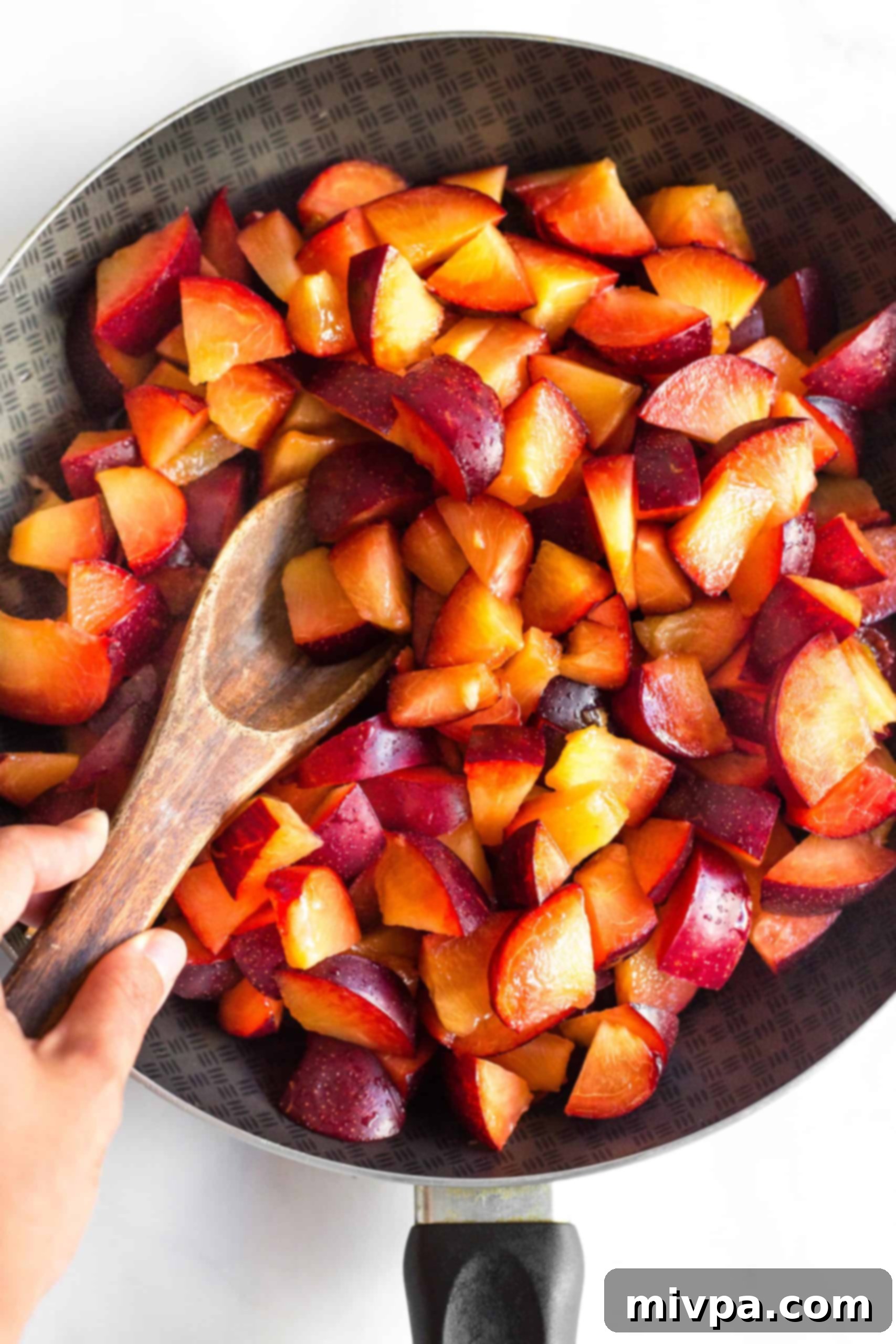 How to Make Plum Jam Without Pectin (Gluten-Free, Vegan) Using a wooden spoon to gently mix cut plum pieces and water in a large non-stick skillet, initiating the jam-making process.