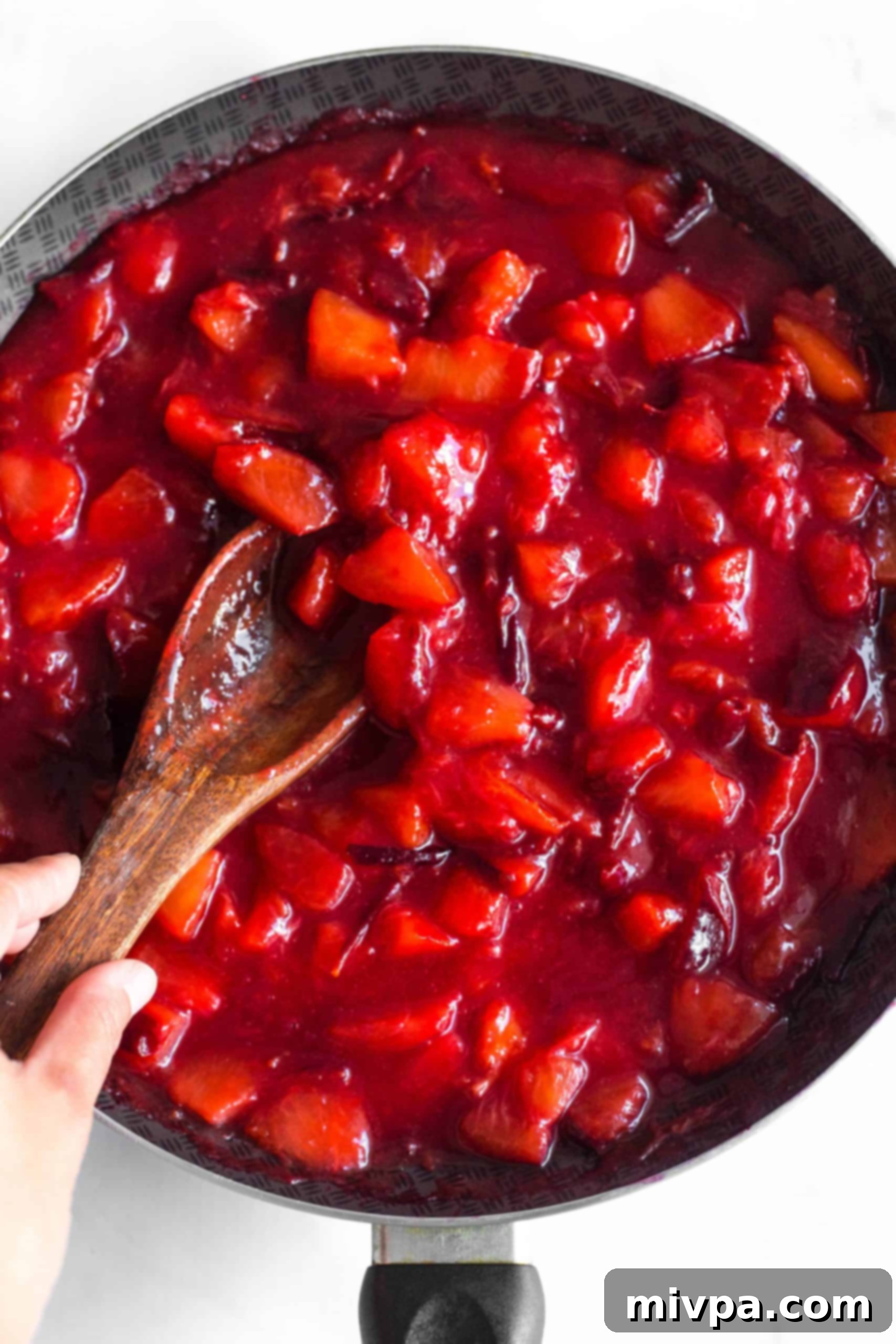Homemade Plum Jam Recipe (Gluten-Free, Vegan, Pectin-Free) A wooden spoon stirring homemade plum jam in a skillet, plums visibly softening and breaking down during the simmering process.