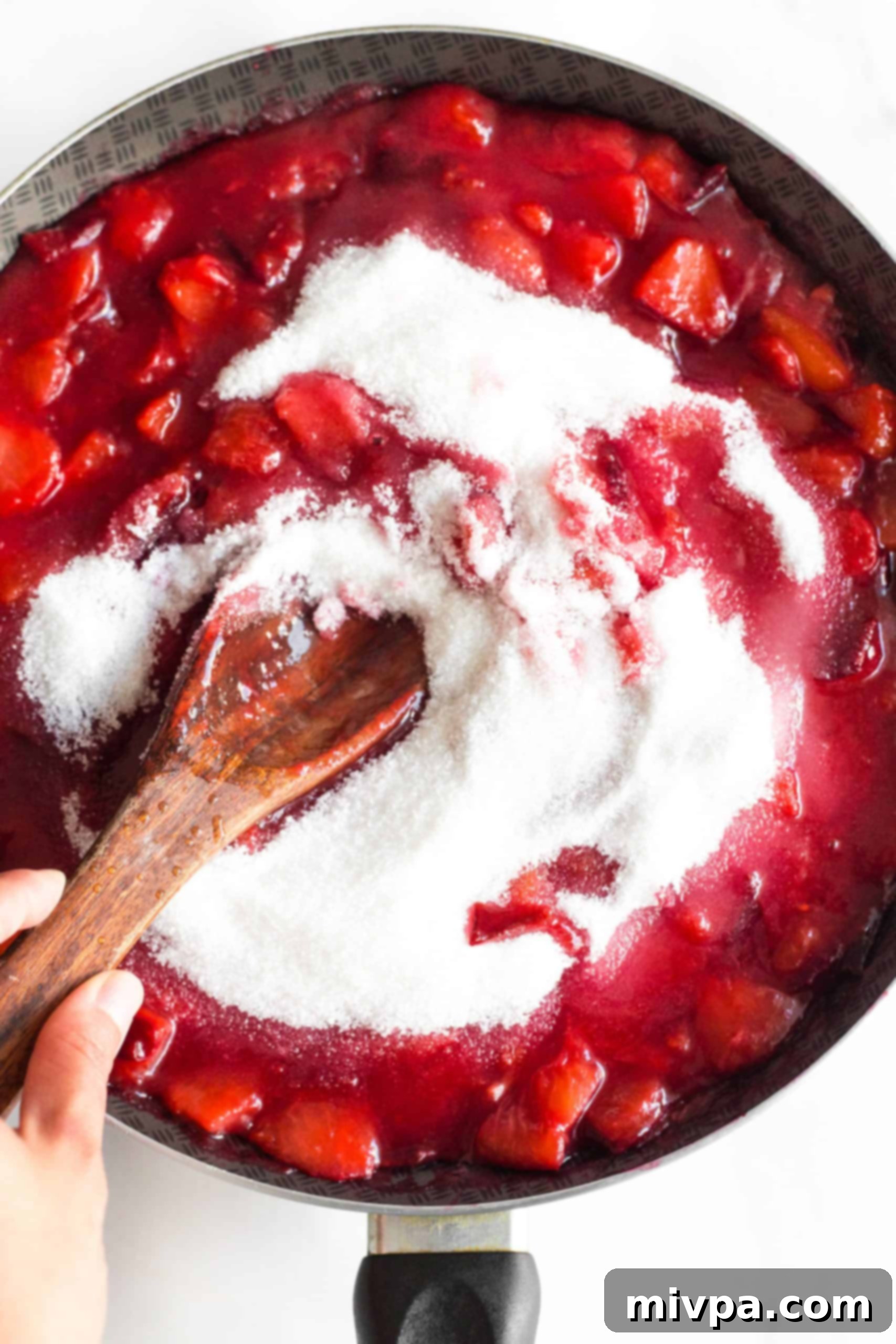 Recipe for Plum Jam (Gluten-Free, Vegan, Pectin-Free) A close-up of a wooden spoon stirring plum jam and fruit in a skillet, as lemon juice and sugar are incorporated into the mixture.