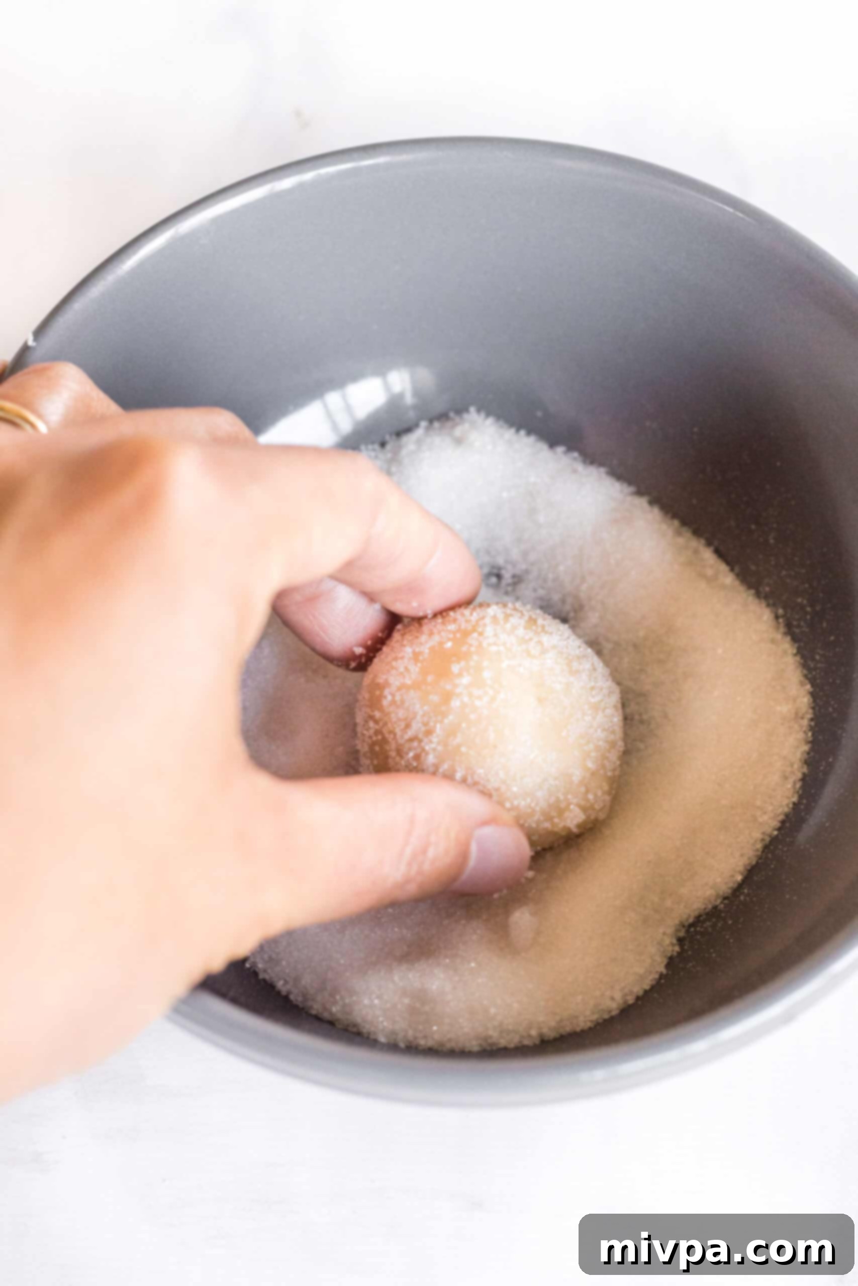 A cookie dough ball being rolled in a bowl of granulated sugar.