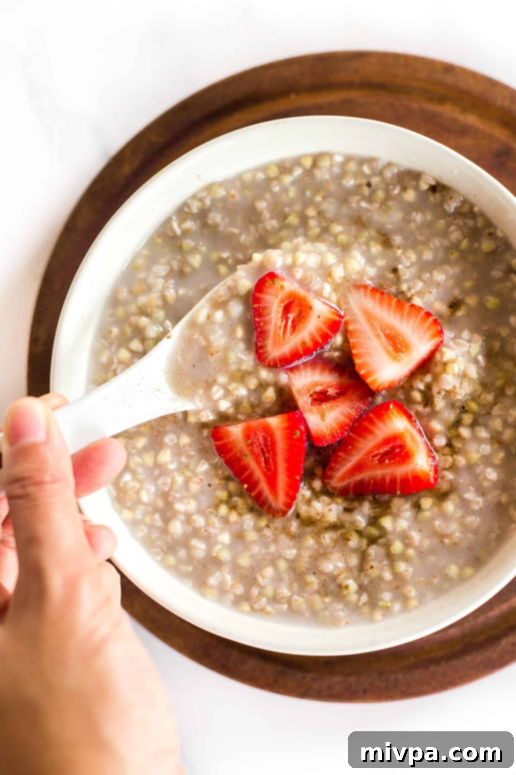 Easy Buckwheat Porridge (Gluten-Free, Vegan) Hand holding a spoon in a bowl of buckwheat porridge with sliced strawberries.