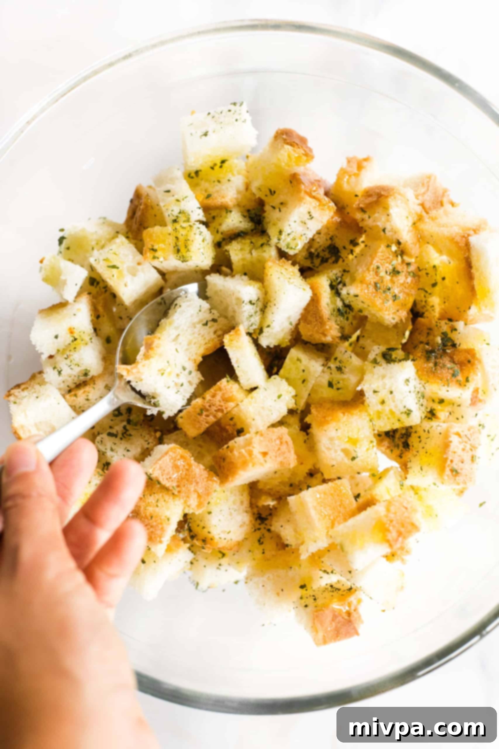 Gluten-free bread cubes being tossed with olive oil and dried herbs in a clear glass mixing bowl.