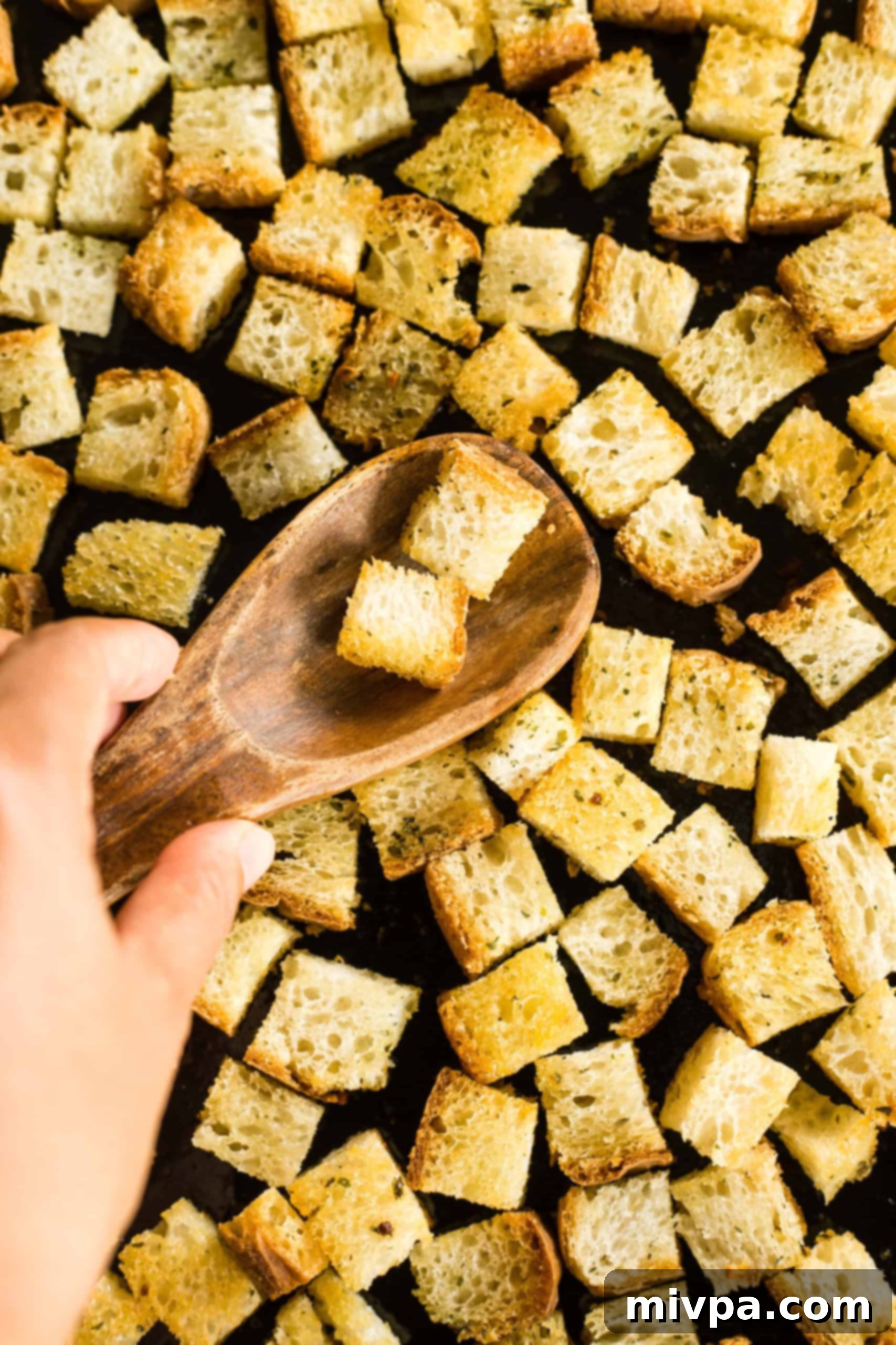 A hand using a wooden spoon to gently toss freshly baked gluten-free croutons on a baking sheet, ensuring even cooling.