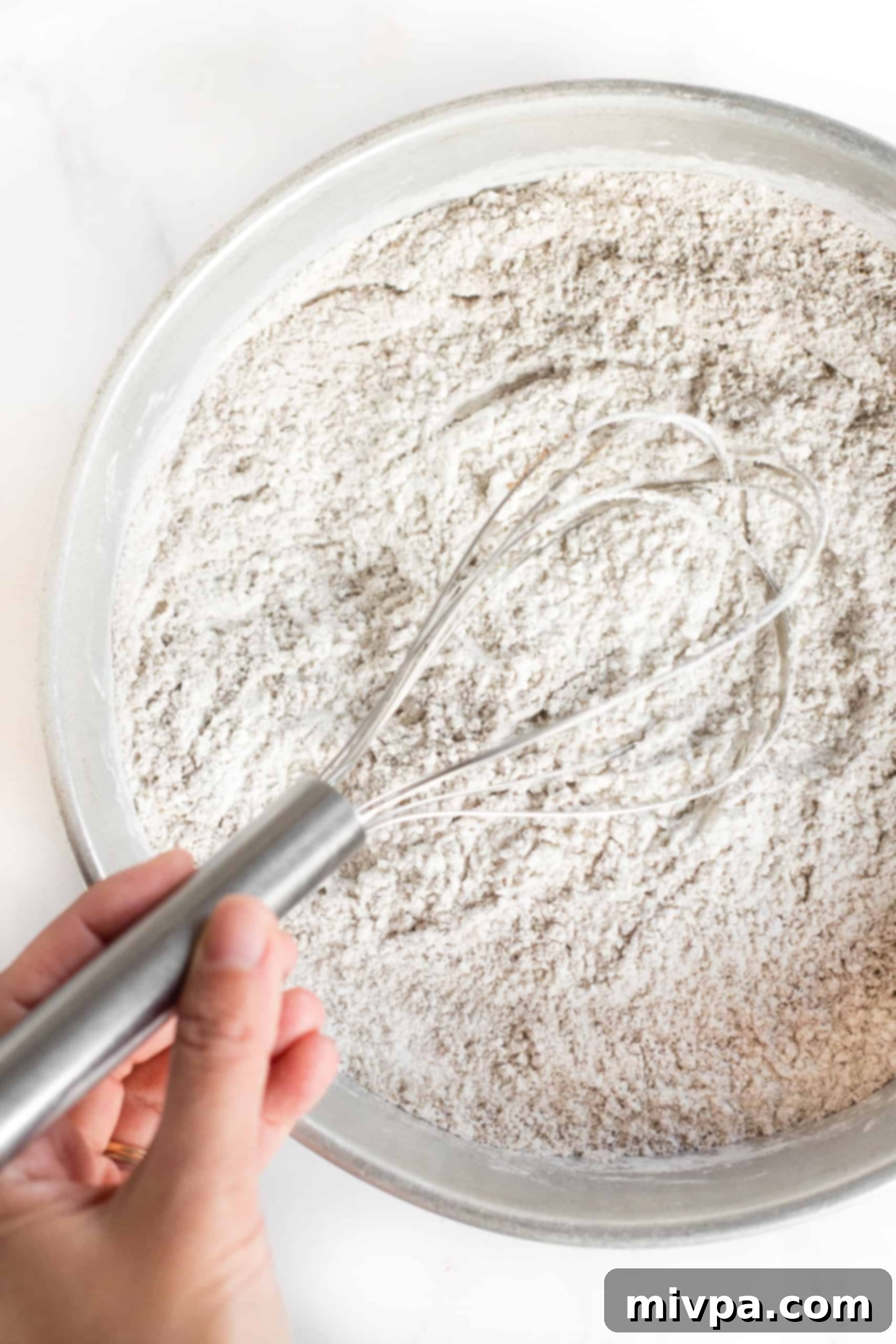 A whisk mixing a light-colored flour mixture in a large metal bowl, ensuring all dry ingredients are combined.