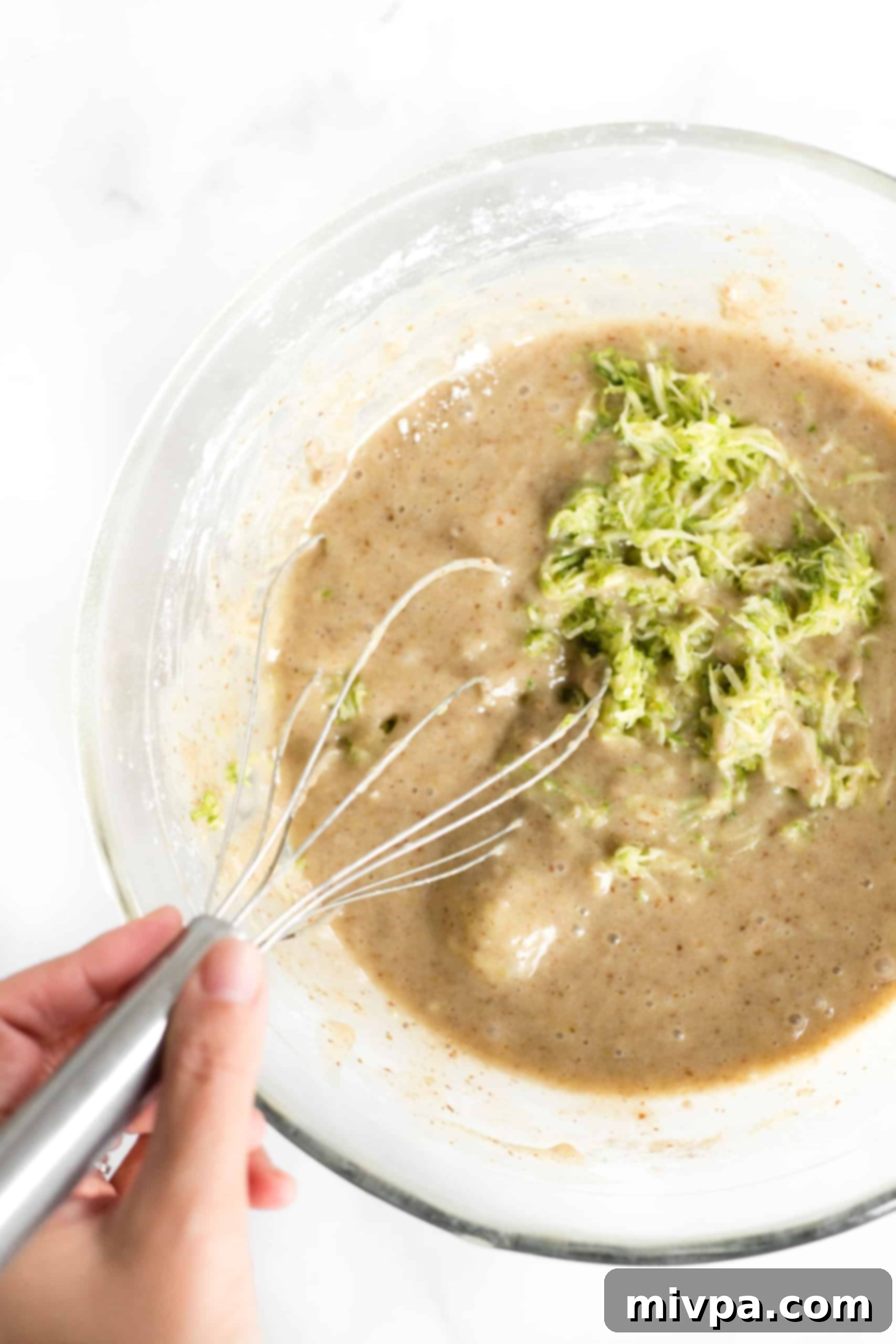 A spatula folding shredded zucchini into the bread batter in a glass bowl, ensuring even distribution.