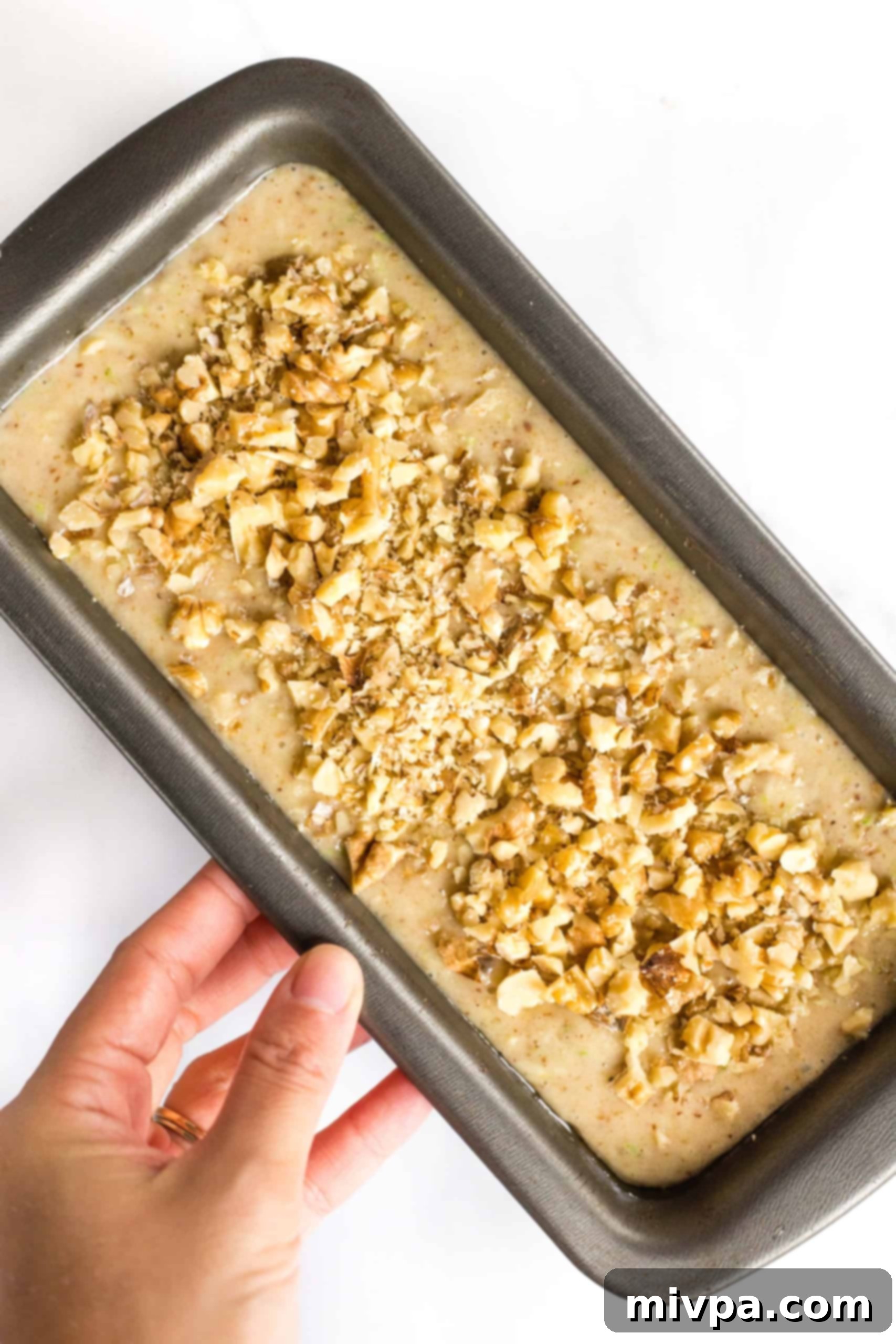 A hand holding a metal loaf pan with zucchini bread batter, liberally sprinkled with chopped walnuts on top.