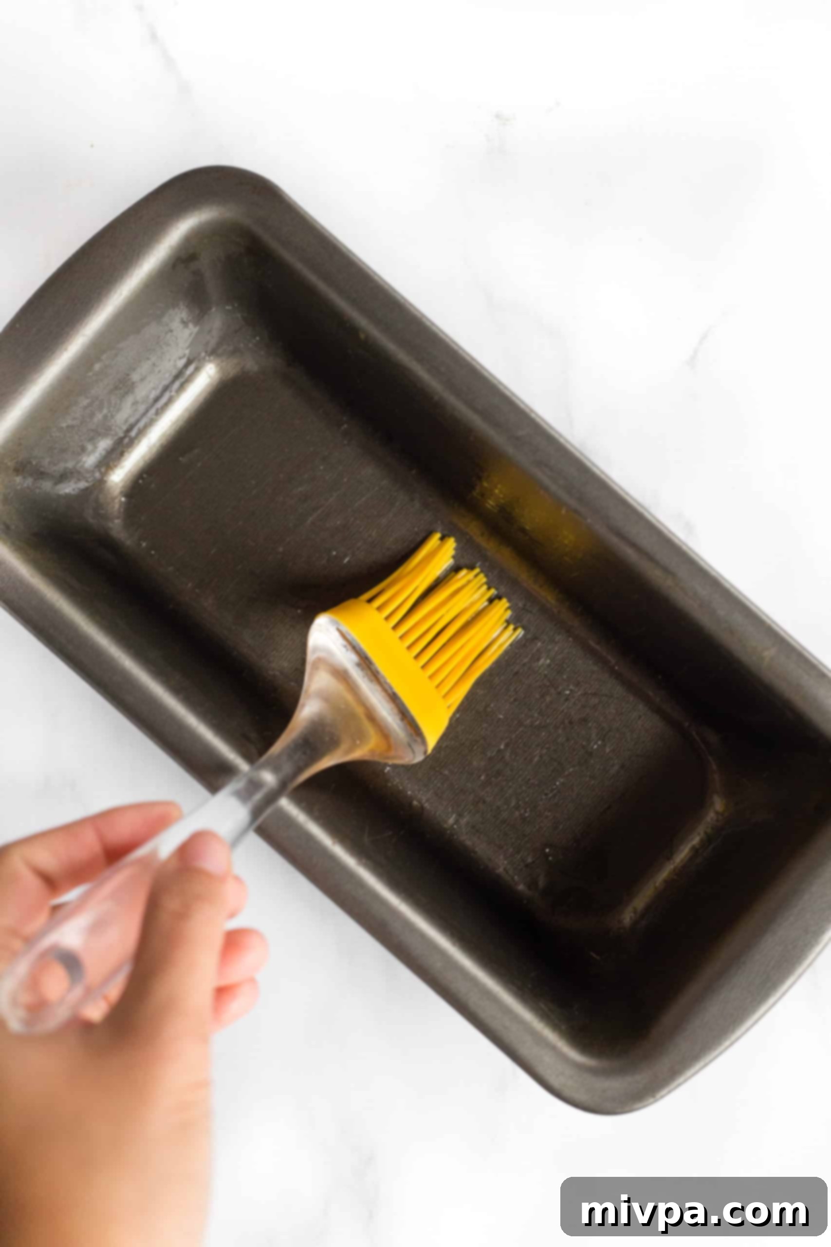 A hand using a silicon brush to grease a metal loaf pan, preparing it for baking.