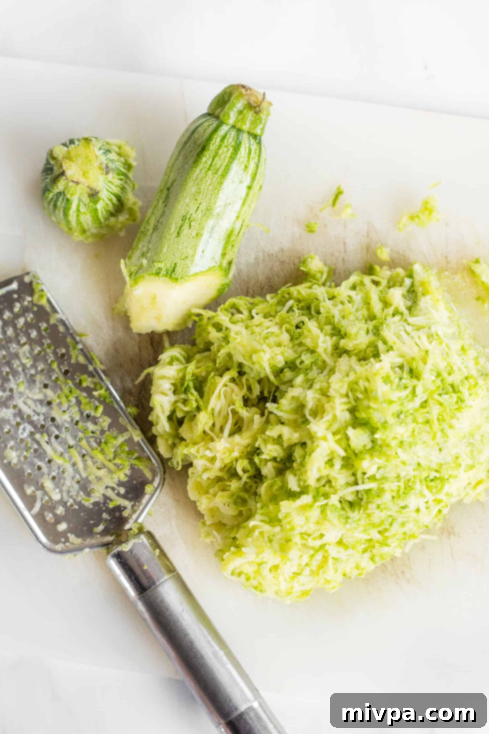 Freshly grated zucchini sitting next to a cheese grater on a wooden chopping board, ready for squeezing.